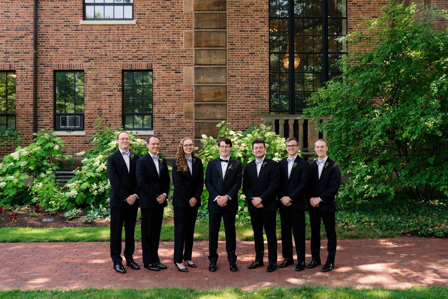 Wedding party in black formal attire posing outdoors in front of a brick building with lush greenery.