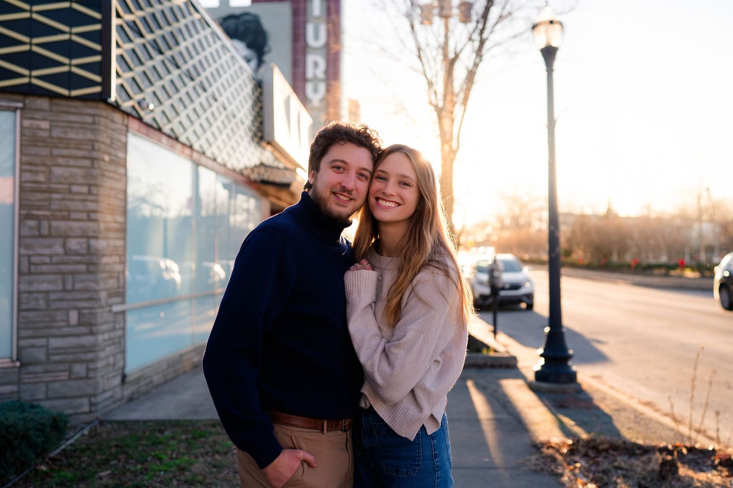 A young couple embraces and smiles on a sunny urban sidewalk at golden hour.