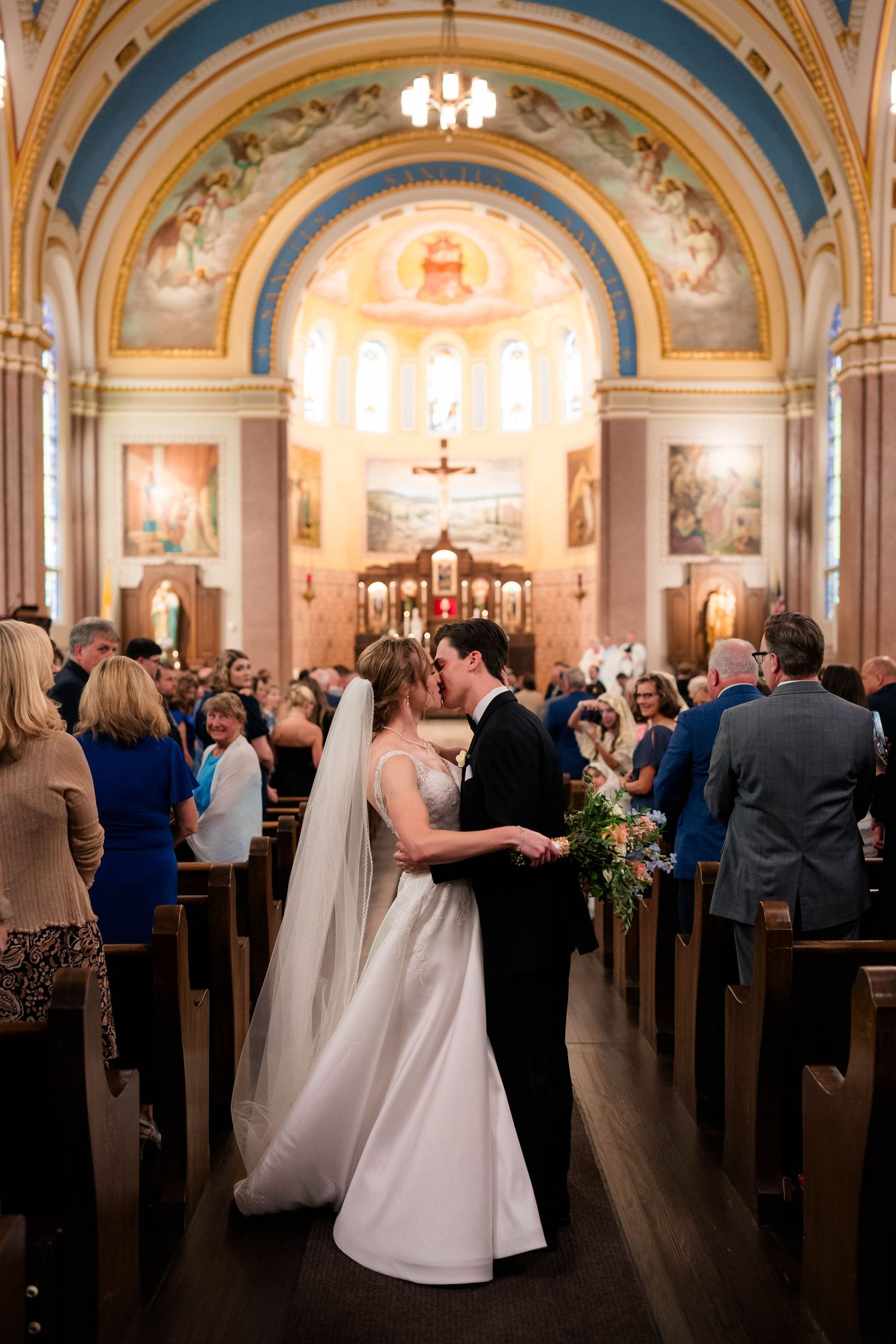 Bride and groom share first kiss in ornate Catholic church aisle with stunning frescoed arched ceiling.