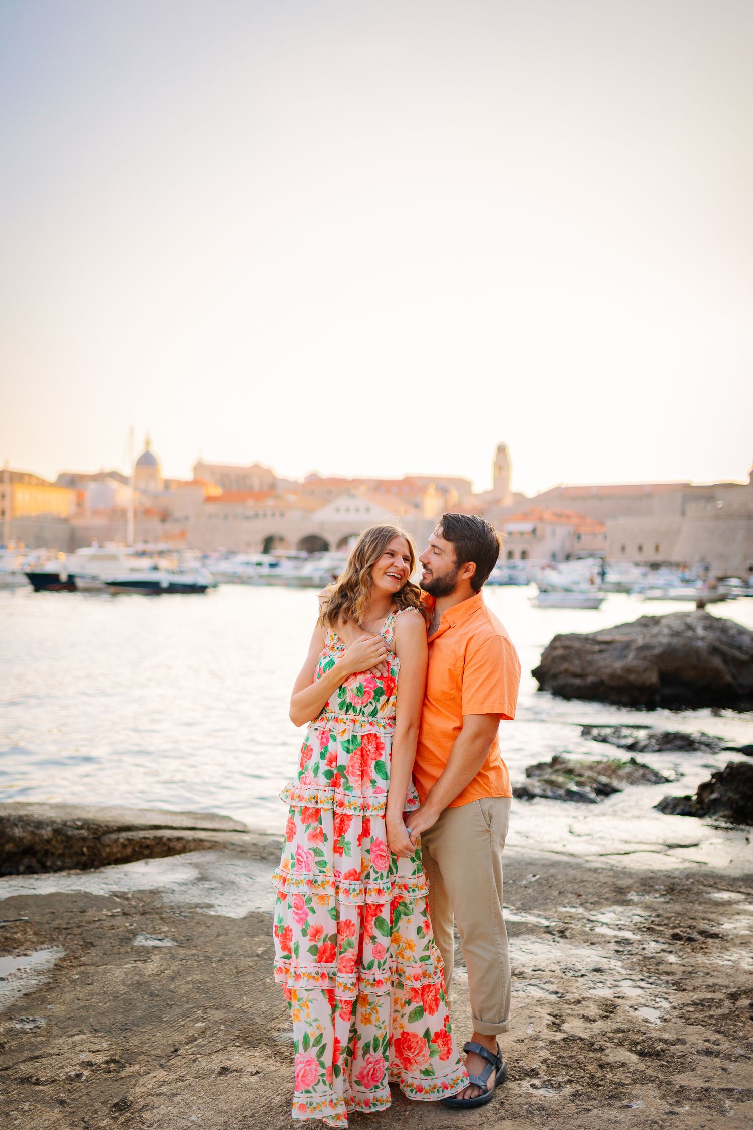Couple embracing at sunset by Dubrovnik harbor in colorful summer outfits.