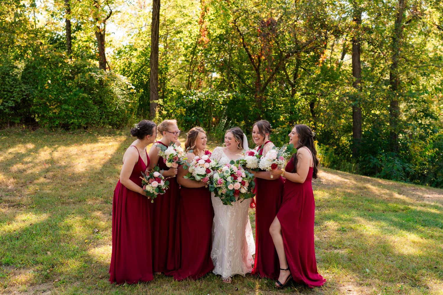 Bride surrounded by five bridesmaids in burgundy gowns holding bouquets in a sunlit forest clearing.