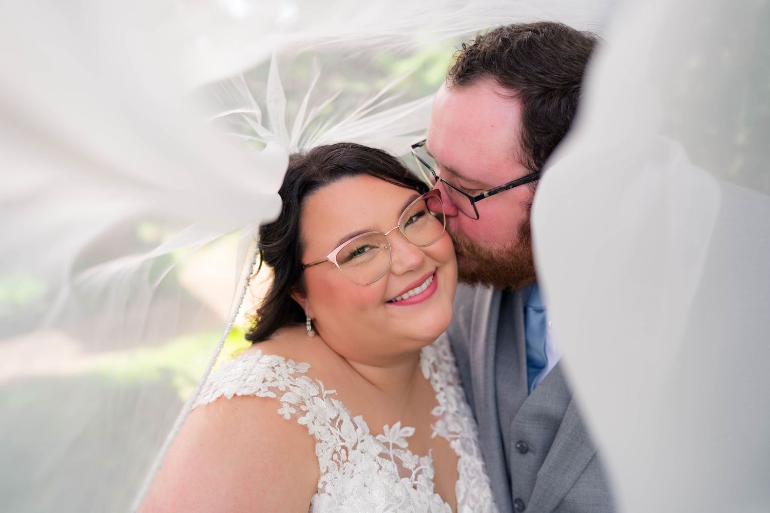 Groom kissing bride on cheek under flowing veil as she smiles warmly at camera.
