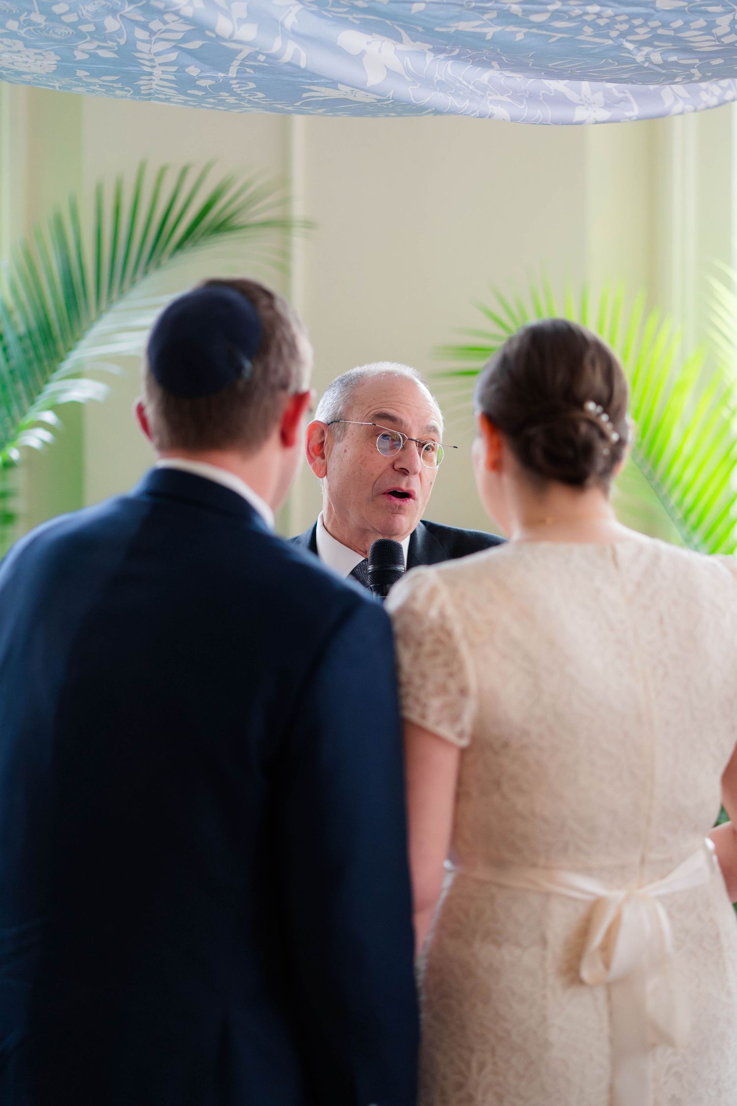 Wedding officiant speaks to bride and groom beneath blue chuppah with palm leaf backdrop.