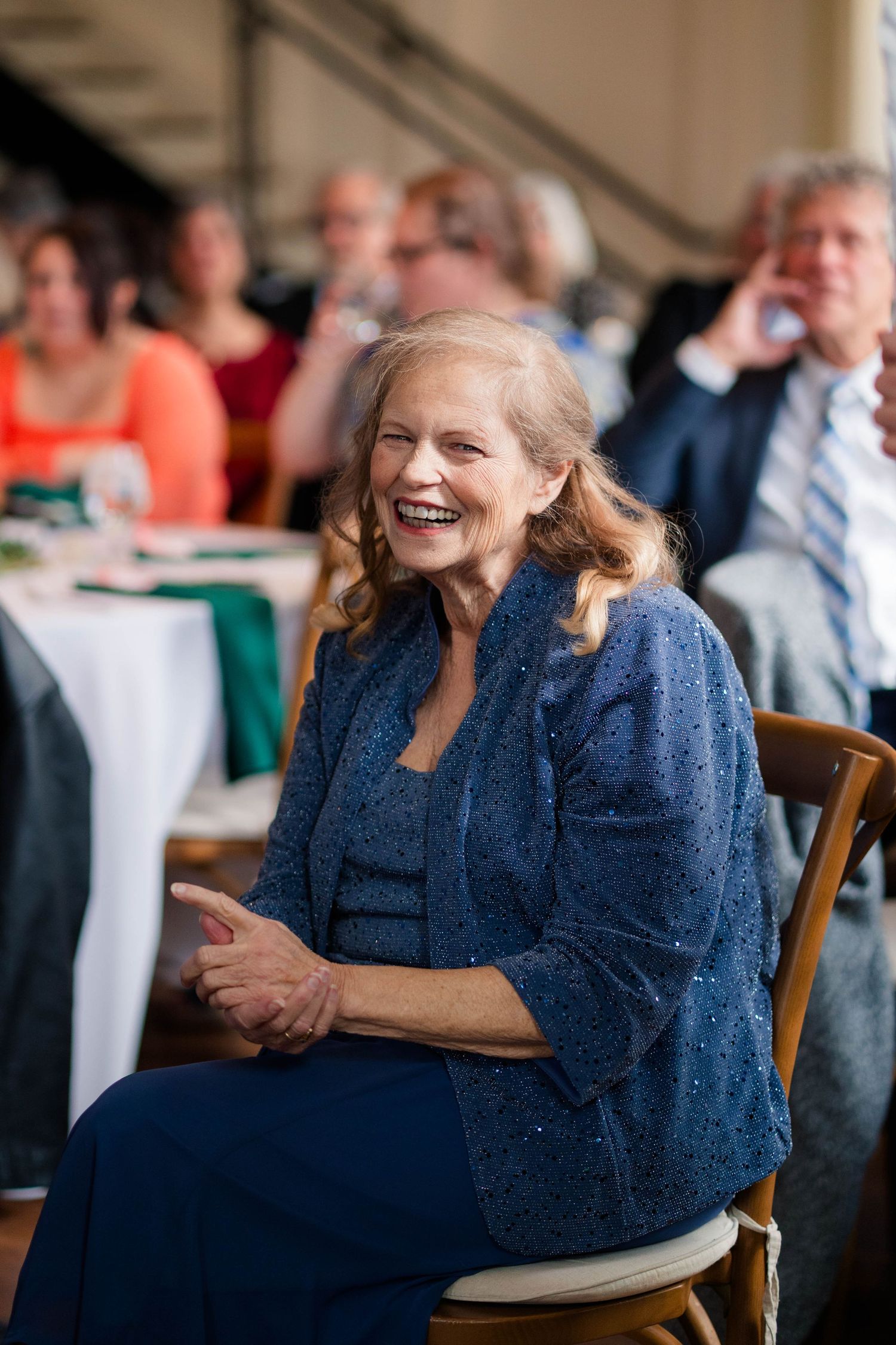 Elegantly dressed older woman laughing joyfully while seated at an outdoor wedding reception.
