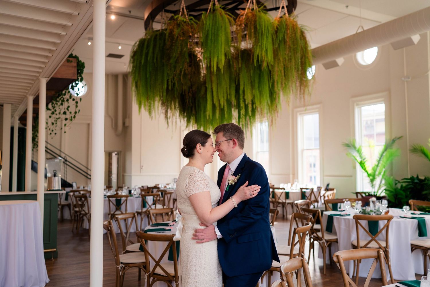 Bride and groom sharing a romantic moment under a cascading fern chandelier in a sunlit wedding reception hall.