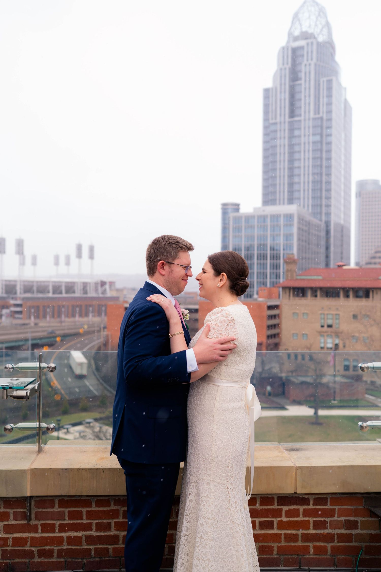 Bride and groom embrace romantically on a rooftop with a misty Cincinnati city skyline in the background.