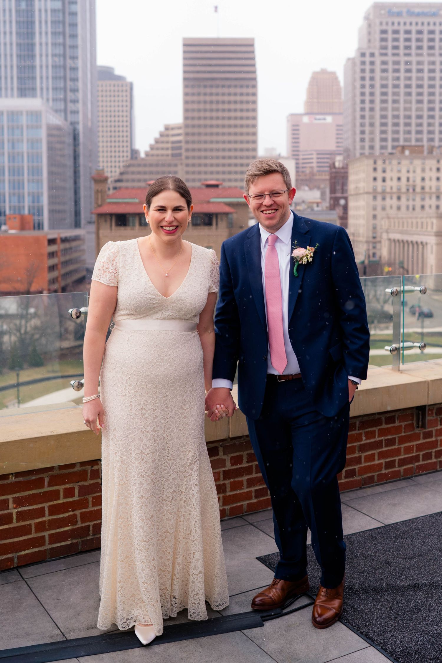 Bride and groom holding hands and smiling on rooftop terrace with urban city skyline behind them.
