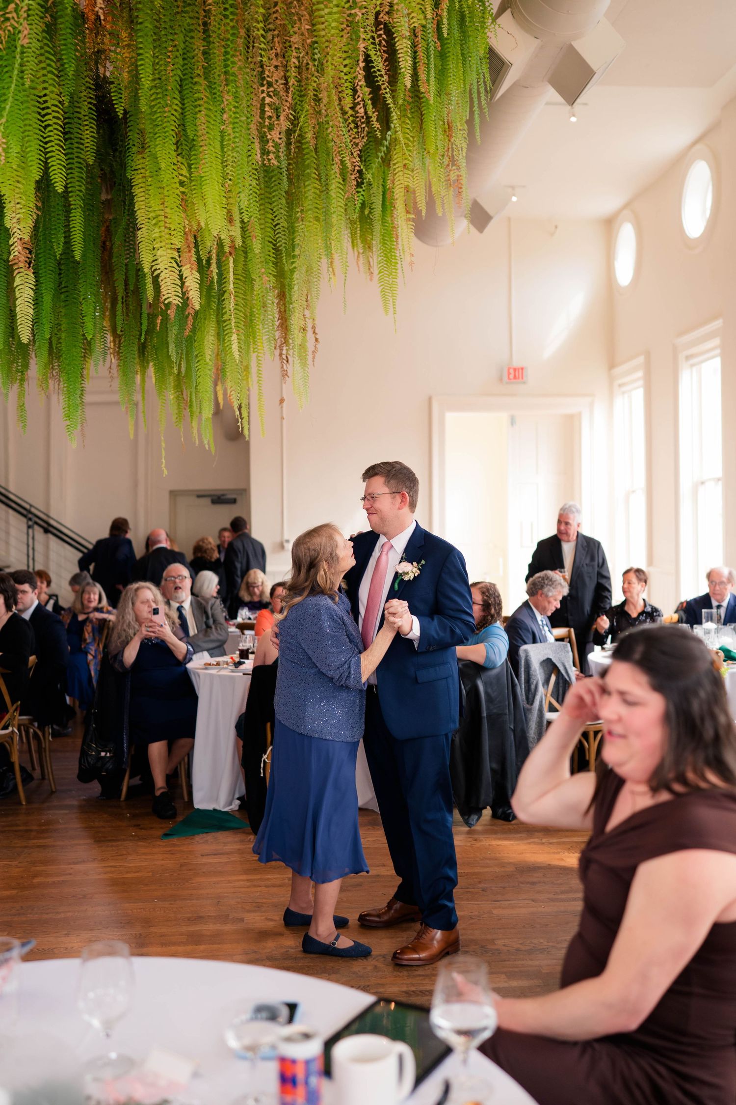 Groom dancing with mother in blue dress at elegant wedding reception with fern decor.