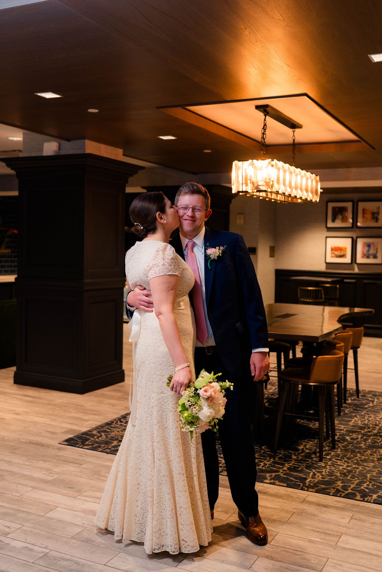 Bride kisses groom on the cheek as he smiles holding a pastel wedding bouquet in a hotel lobby.