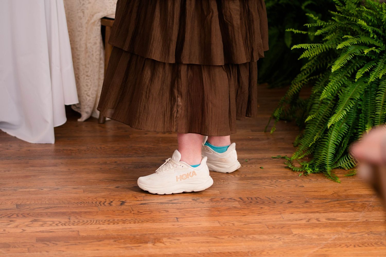 Close-up of white sneakers with blue accents worn under a brown ruffled dress on hardwood floor.