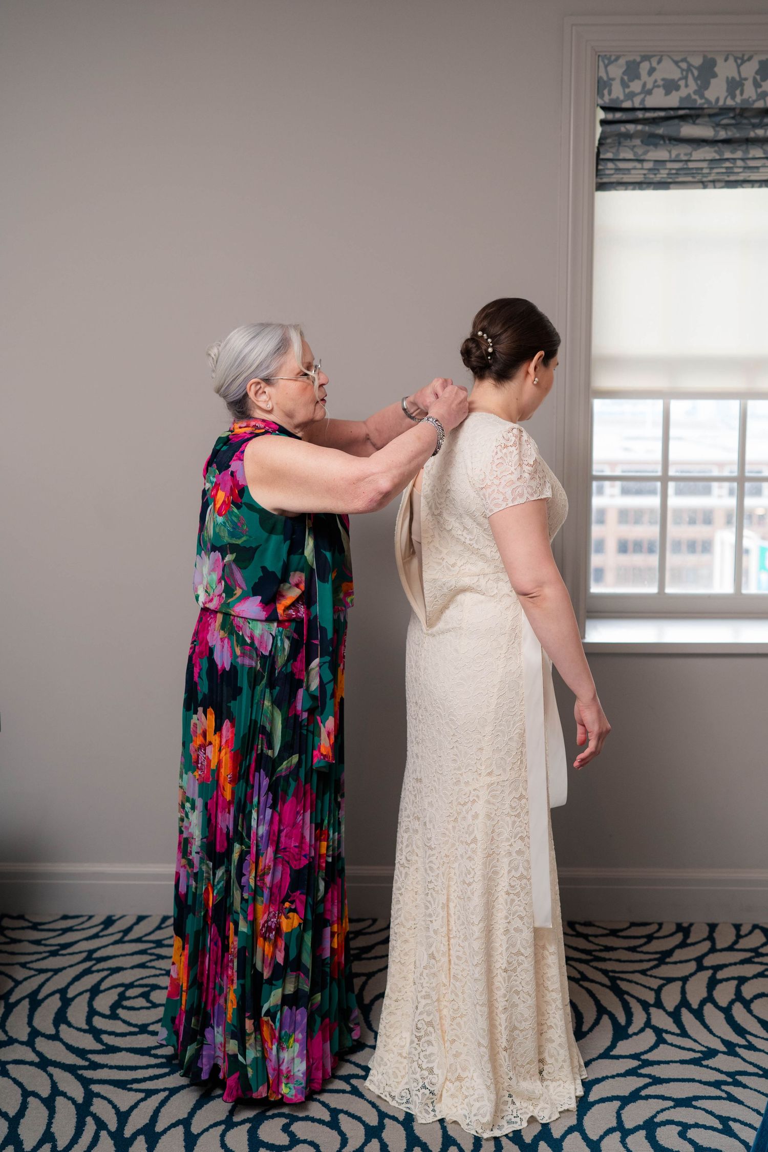 Mother in colorful floral gown fastens the back of bride's lace wedding dress.