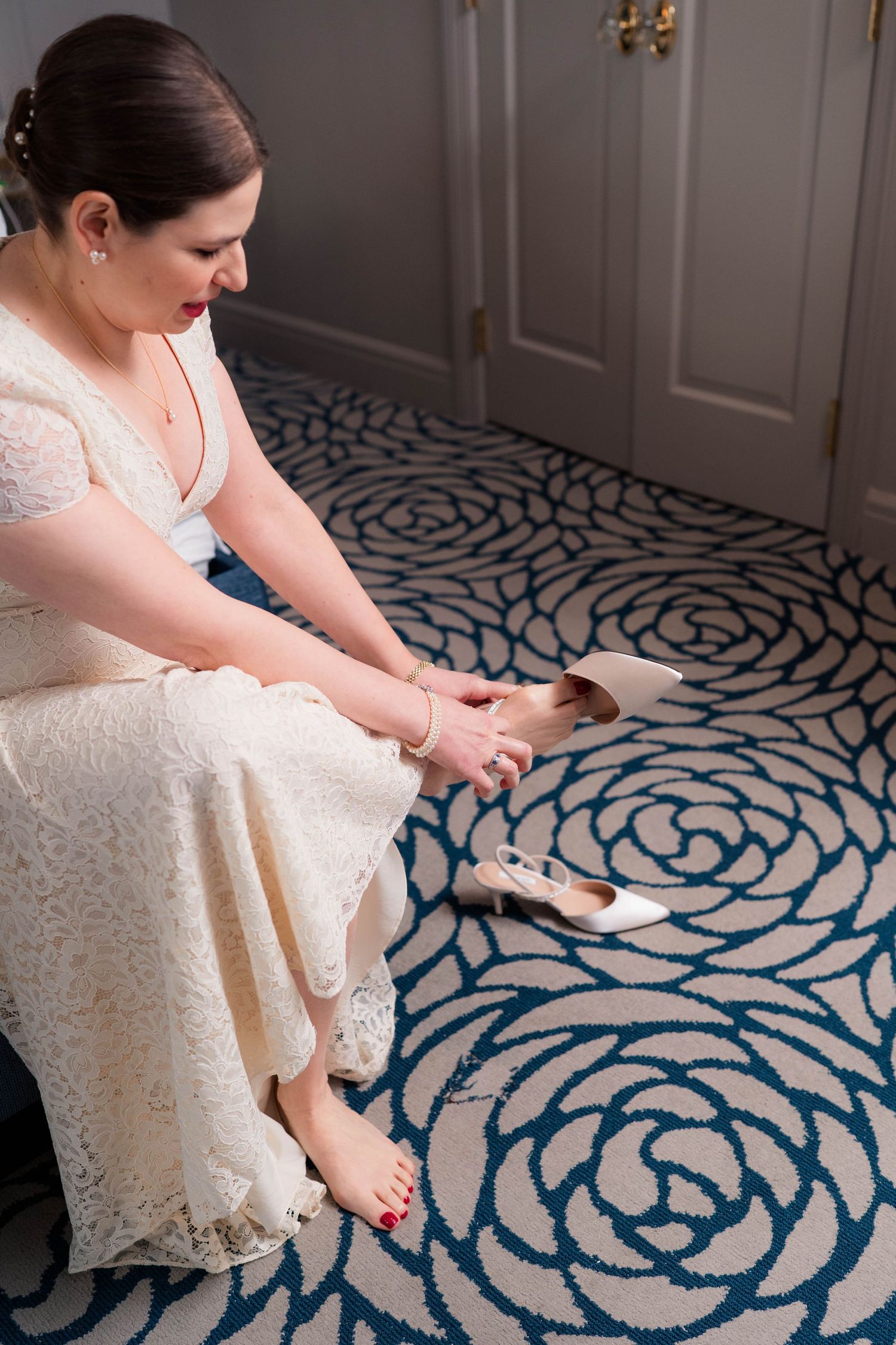 Bride in lace wedding dress sits on floral carpet putting on ivory heels.