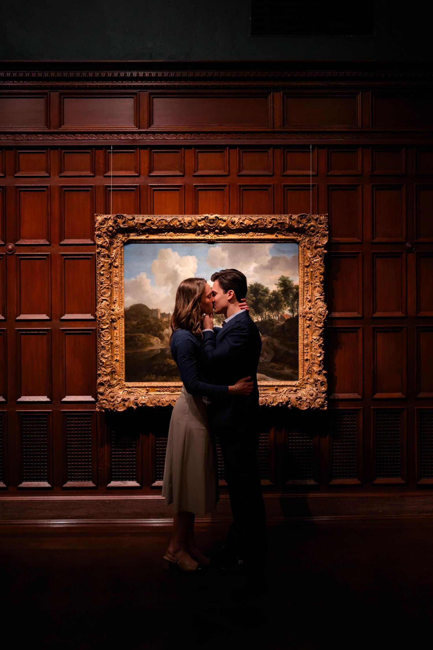 Romantic couple sharing a kiss before a gilded painting in a dark wood-paneled gallery.