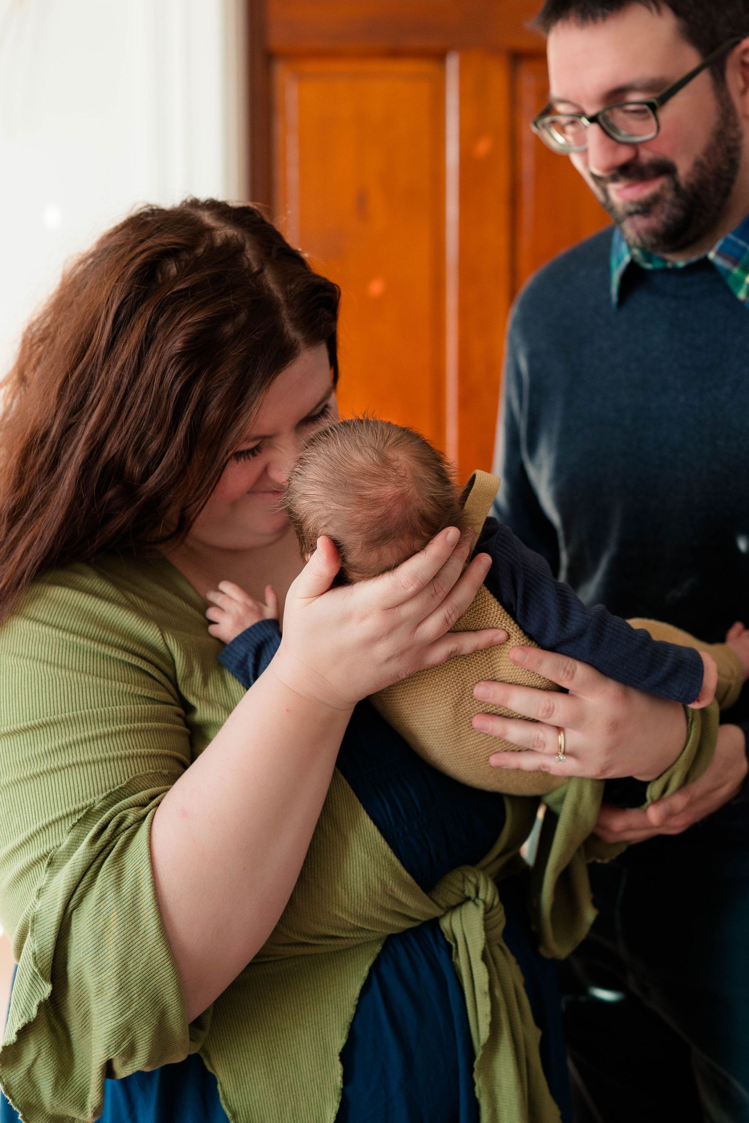 New mother in green dress tenderly kissing newborn baby while smiling father watches nearby.