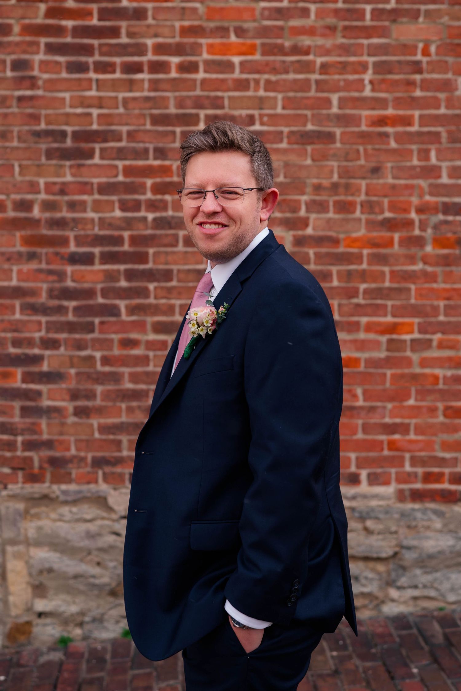A smiling groom in a navy suit with boutonniere poses against a rustic red brick wall.