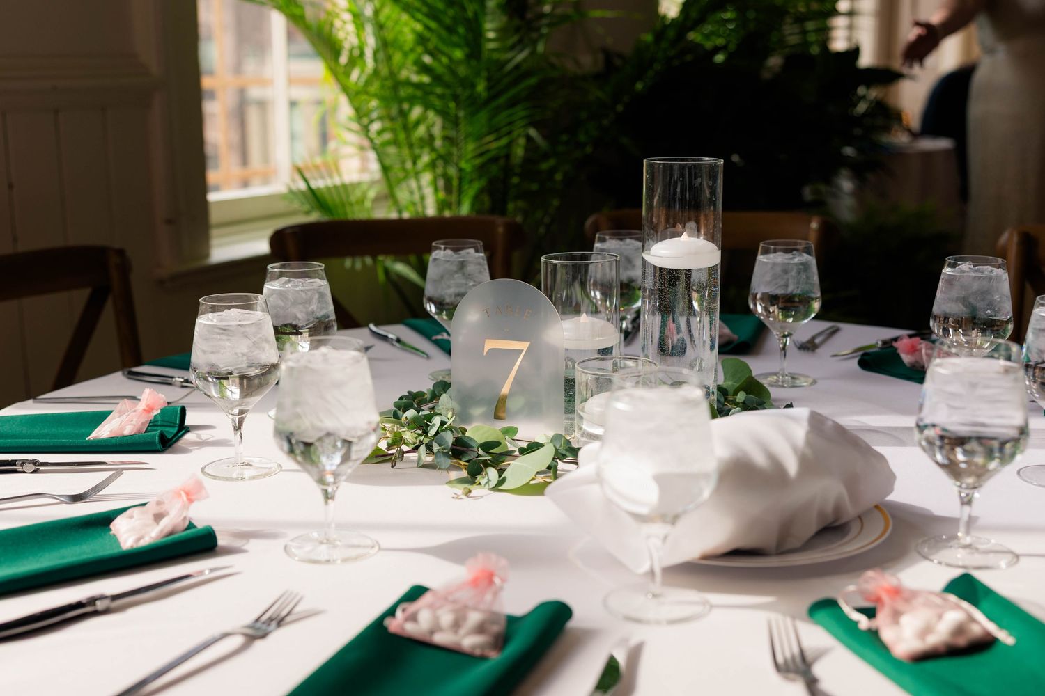 Elegant wedding reception table with green napkins, white linens, crystal glassware, and a floral centerpiece with table number 7.