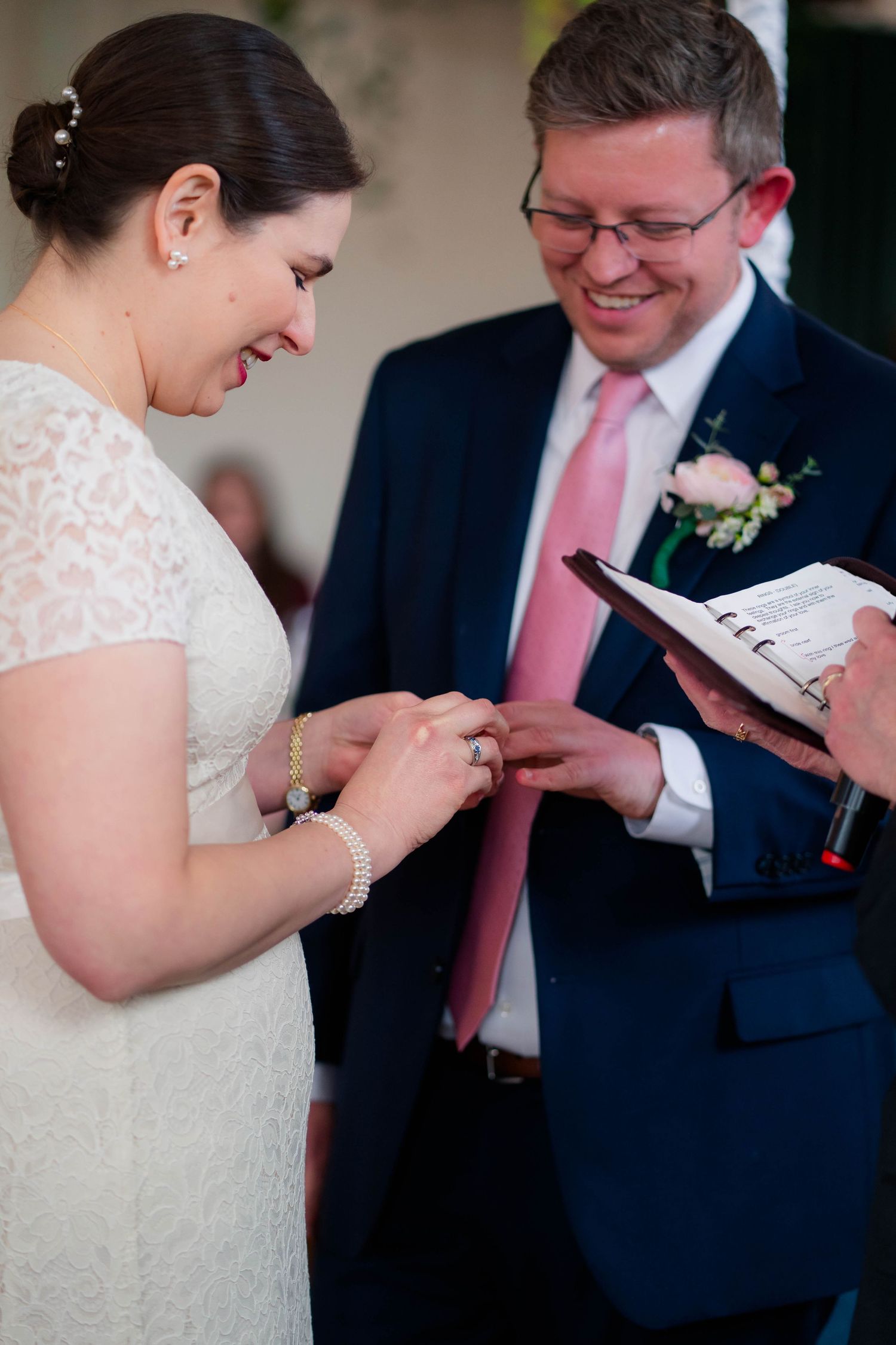 Bride places wedding ring on finger while groom smiles during ring exchange ceremony.