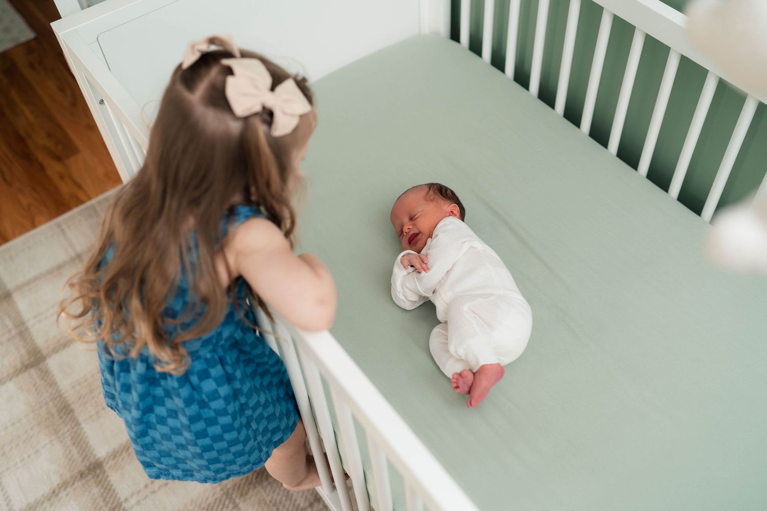 Young girl in blue dress looks over crib railing at sleeping newborn sibling.
