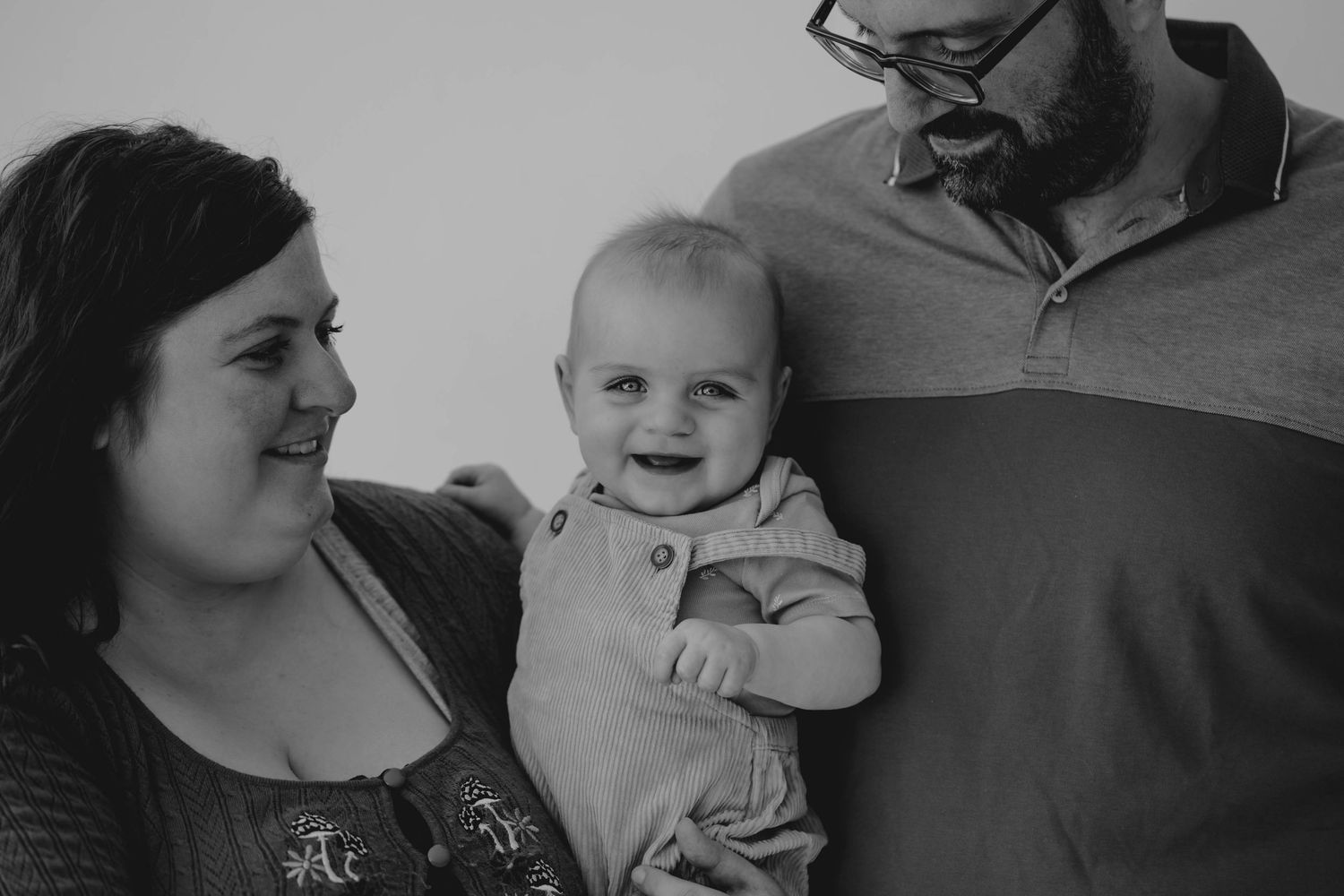 Black and white portrait of smiling baby held by parents in casual family photo.