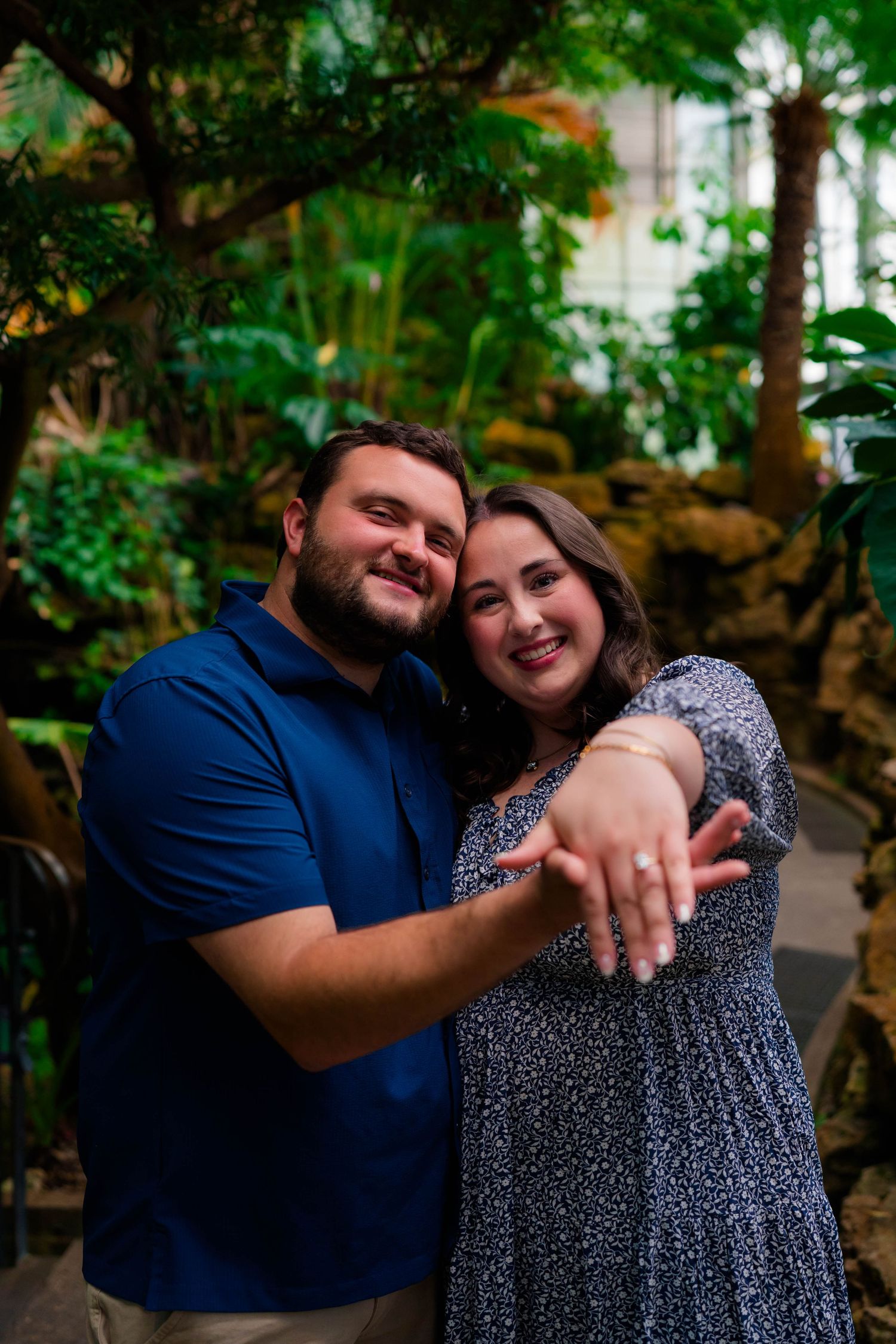Joyful newly engaged couple showing off engagement ring while hugging in a tropical botanical garden.