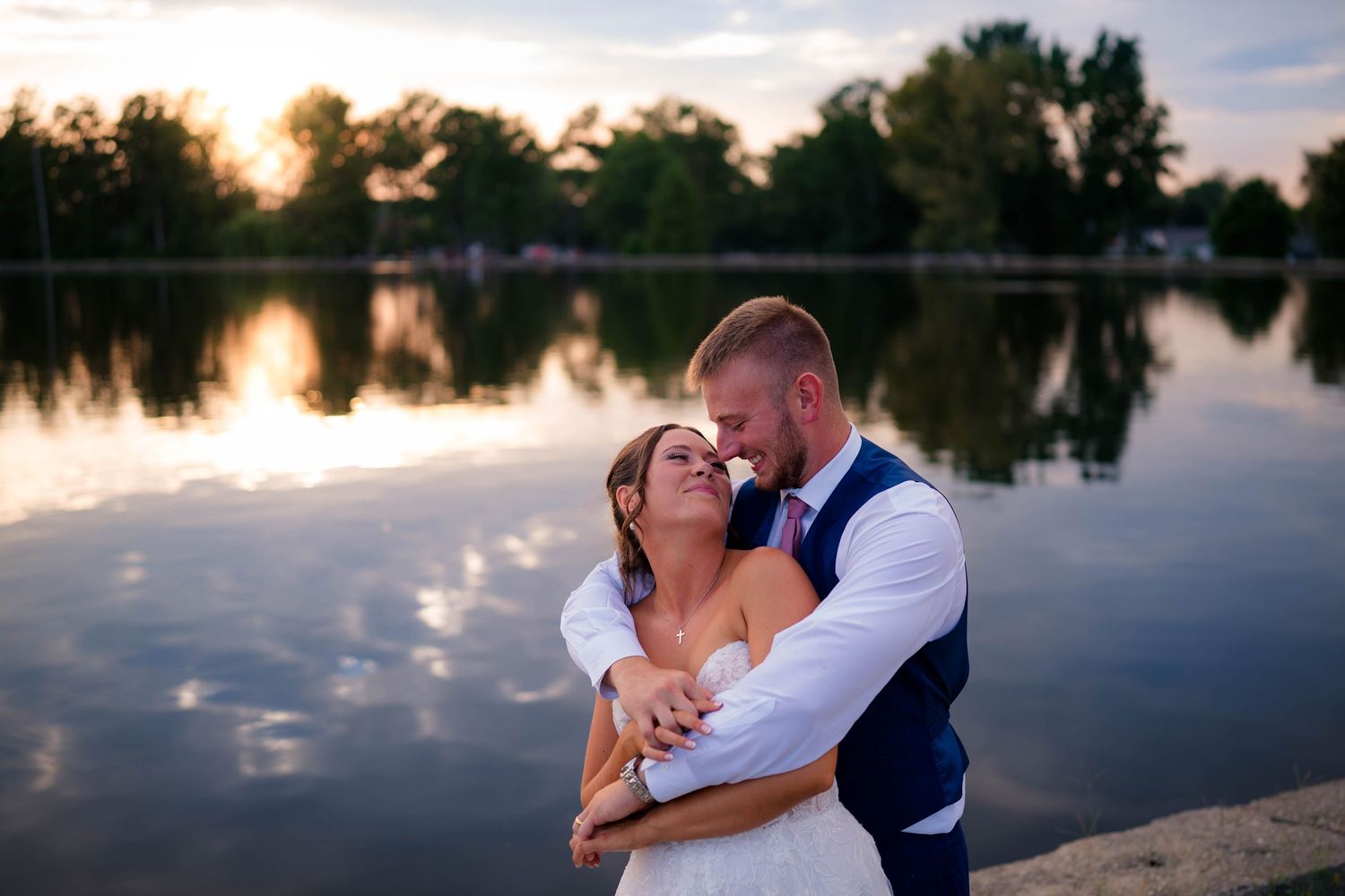Bride and groom share a romantic embrace by a reflective lake at sunset, groom in navy vest, bride in white gown.
