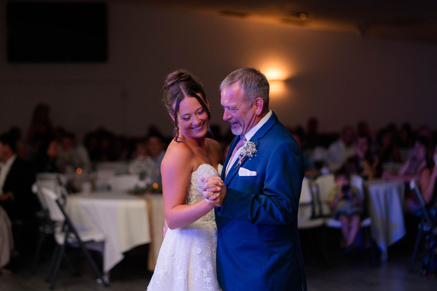 Bride and father share an emotional first dance at wedding reception in elegant venue with guests seated in background.
