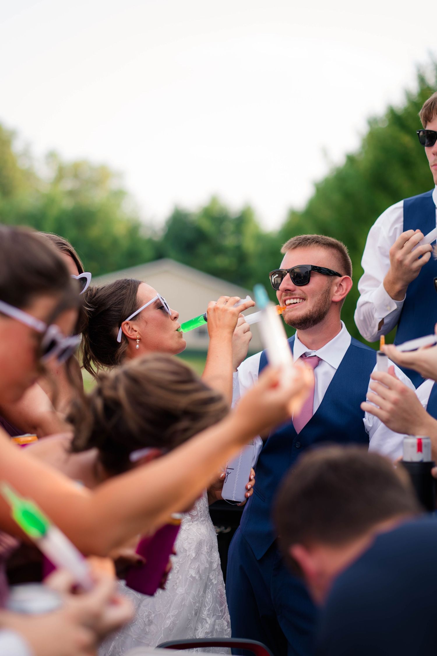 Smiling groom in navy vest watches bride drink at lively outdoor wedding reception with guests.