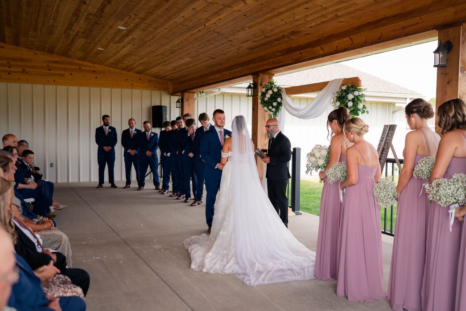 Bride and groom exchange vows at floral arch with wedding party in navy and dusty rose attire.