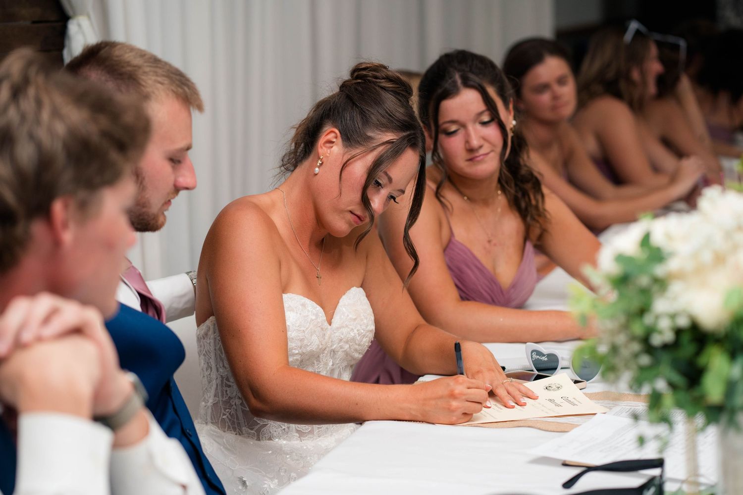 Emotional bride wiping tears while signing wedding documents at reception table with bridesmaids nearby.