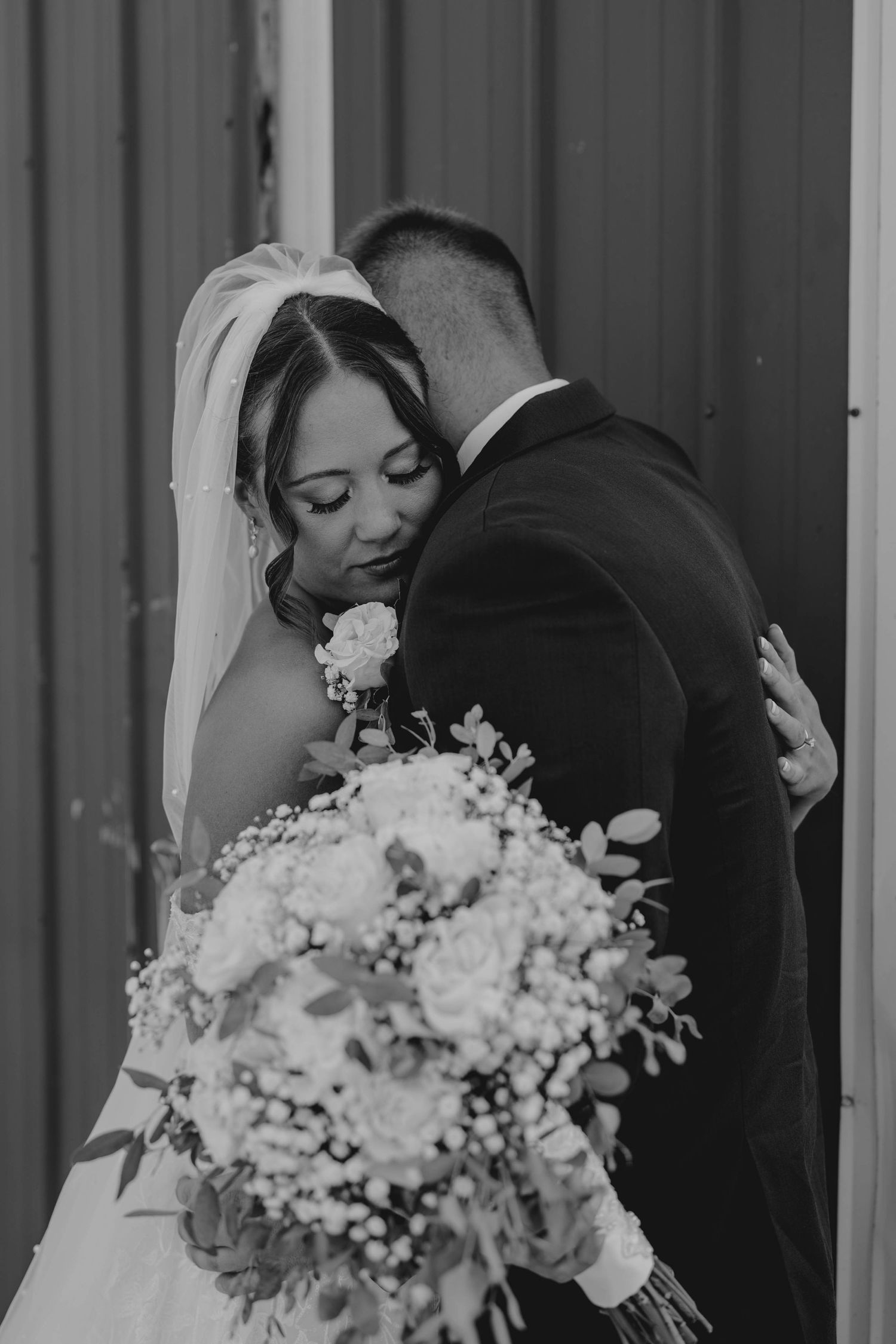 Black and white photo of bride and groom sharing tender embrace, bride holding white floral bouquet.
