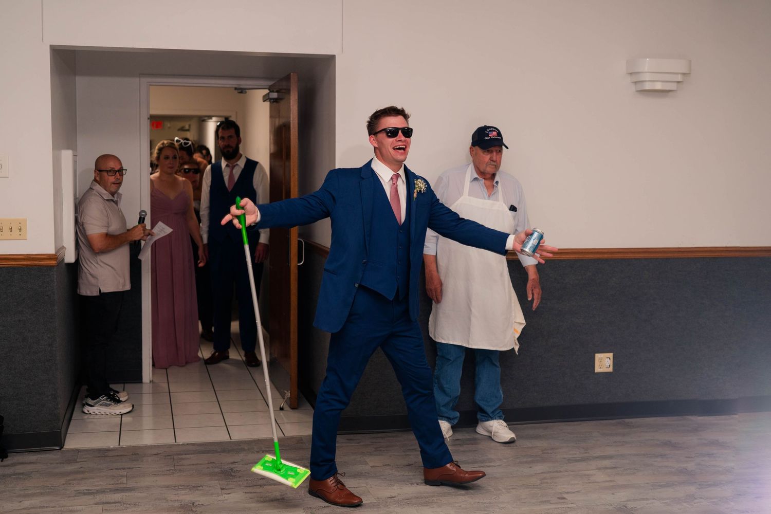 Groomsman in navy suit humorously sweeps dance floor with broom at wedding reception entrance.