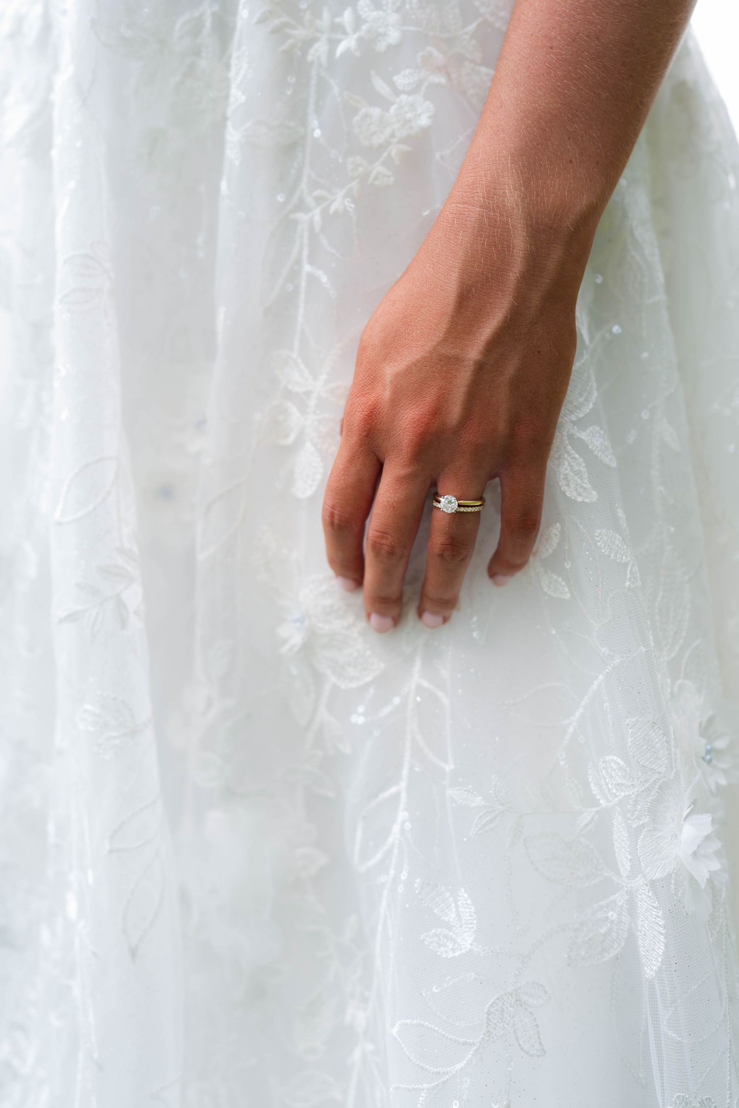 Close-up of bride's hand with diamond engagement ring resting on white lace wedding dress.