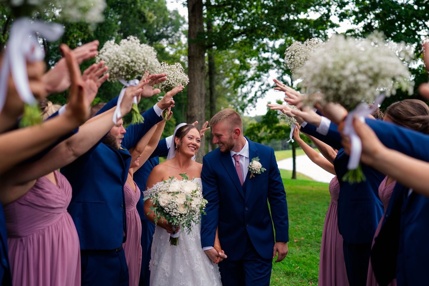 Bride and groom walk through arch of raised bouquets held by wedding party in navy and mauve attire.