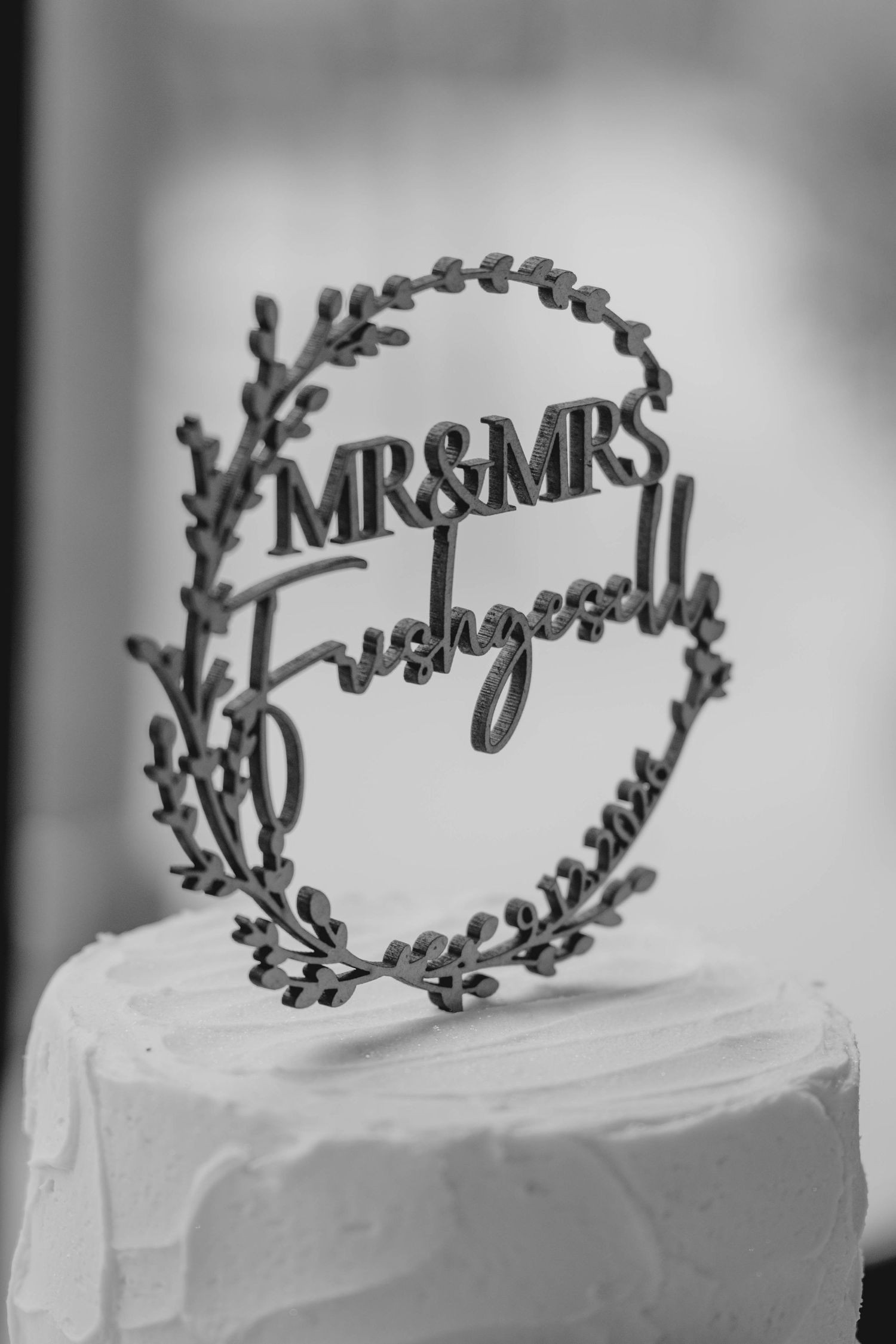 Black and white photo of Mr and Mrs wedding cake topper with wreath design on white cake.