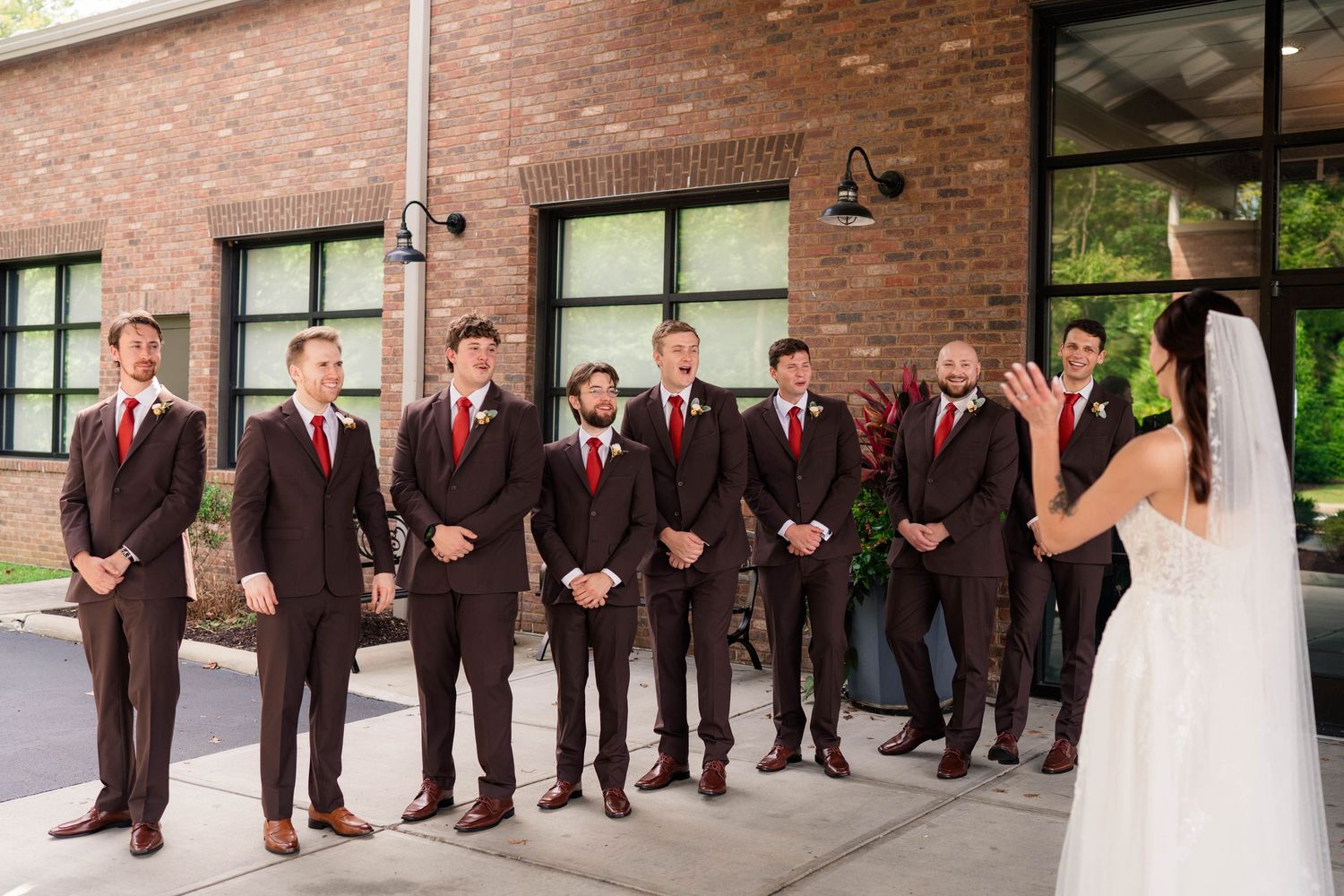Bride reveals her wedding dress to groomsmen in dark suits with red ties outside a brick building.