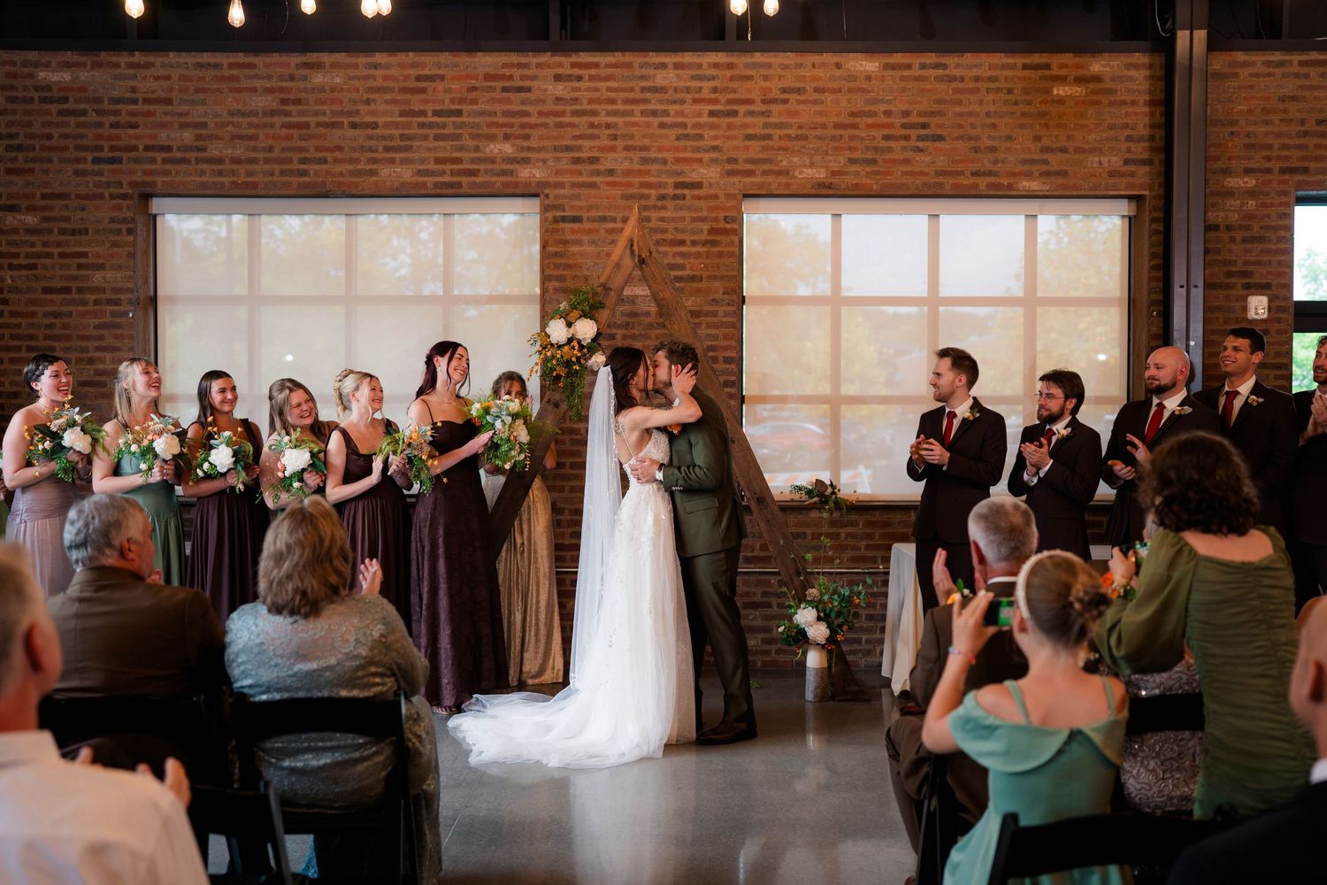 Bride and groom share first kiss as wedding party and guests applaud at indoor brick venue ceremony.