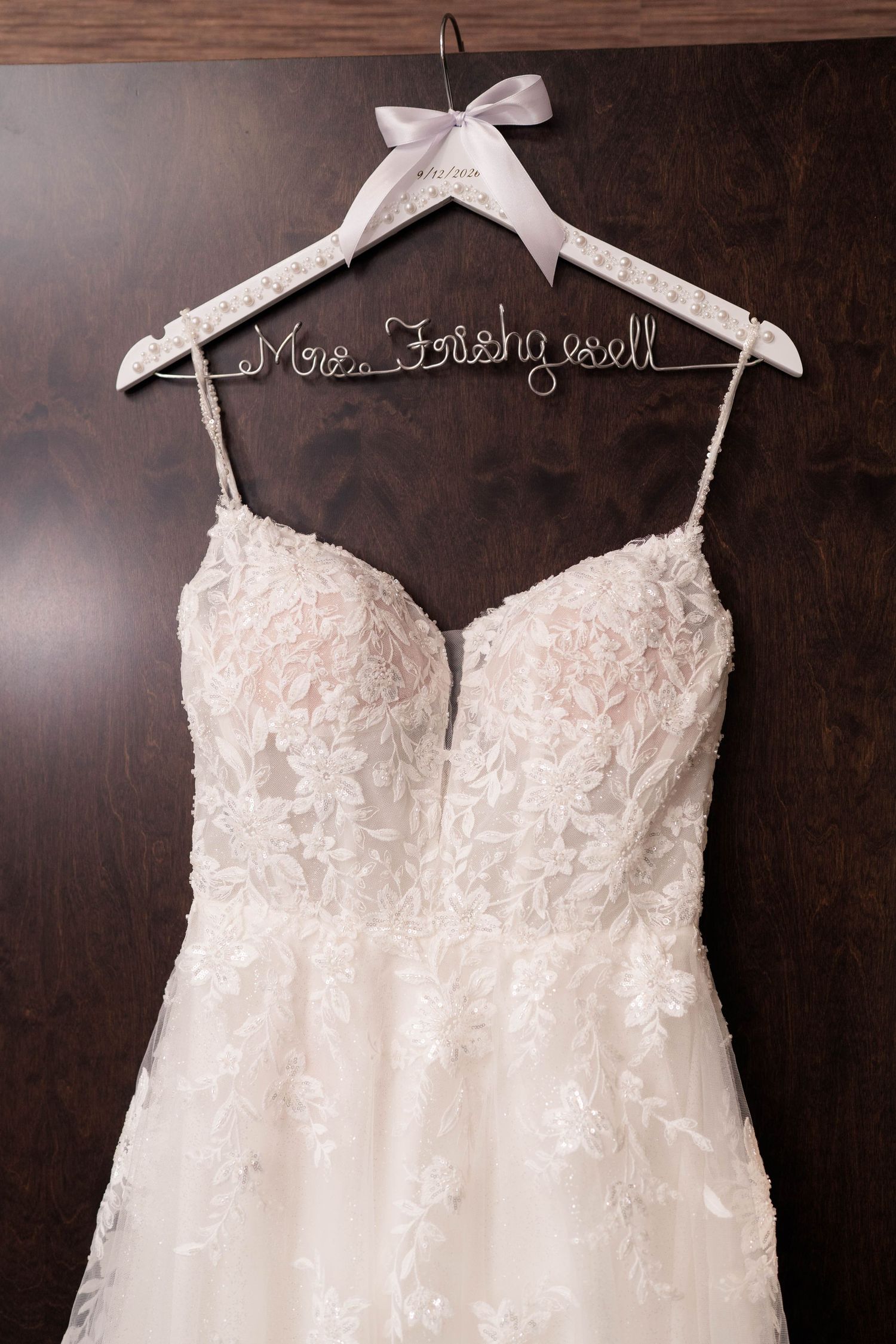 Elegant lace wedding dress on a custom wire name hanger reading Mrs. Fishywell against a dark wood background.