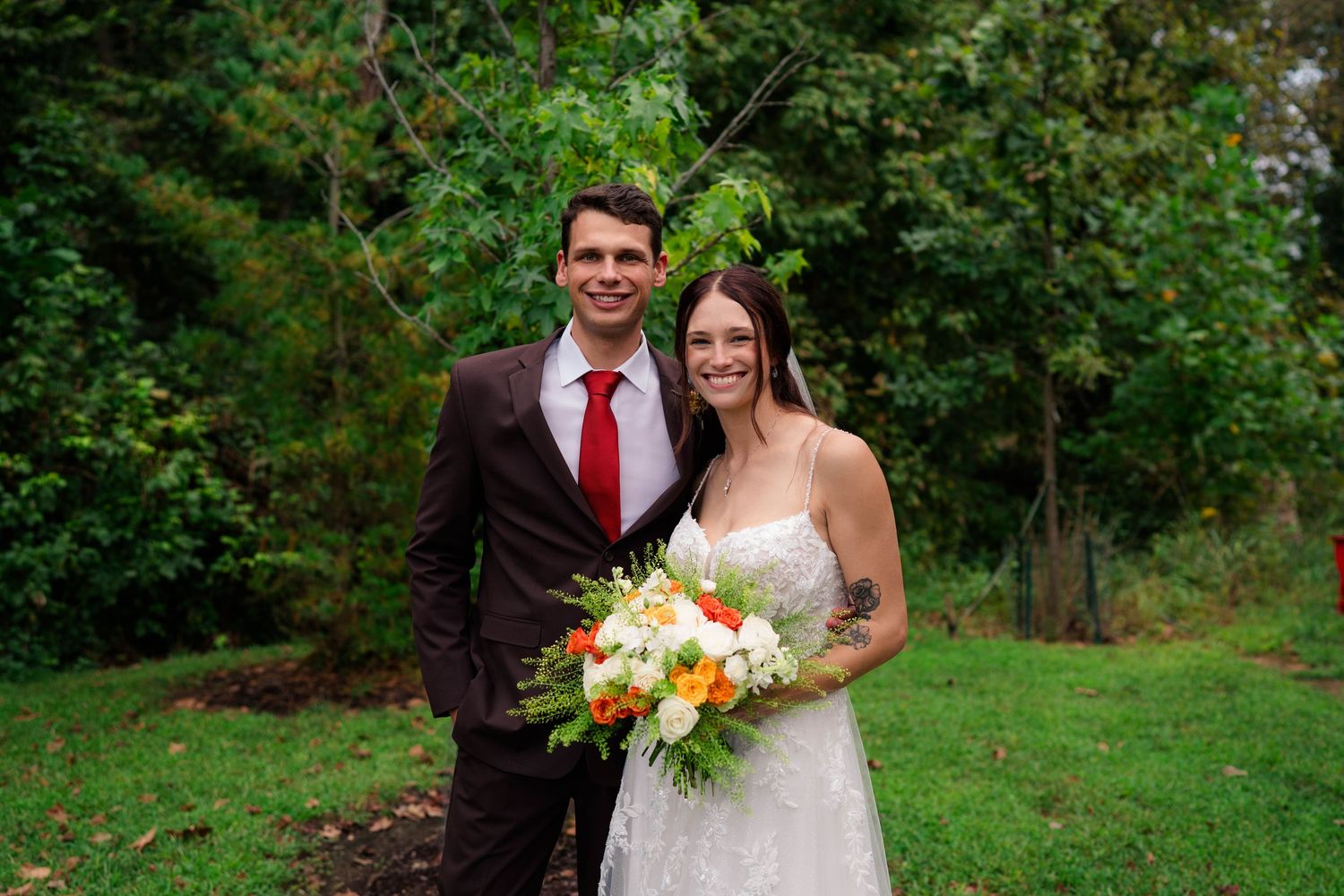 Smiling bride and groomsman pose together outdoors, bride holding vibrant orange and white bouquet.