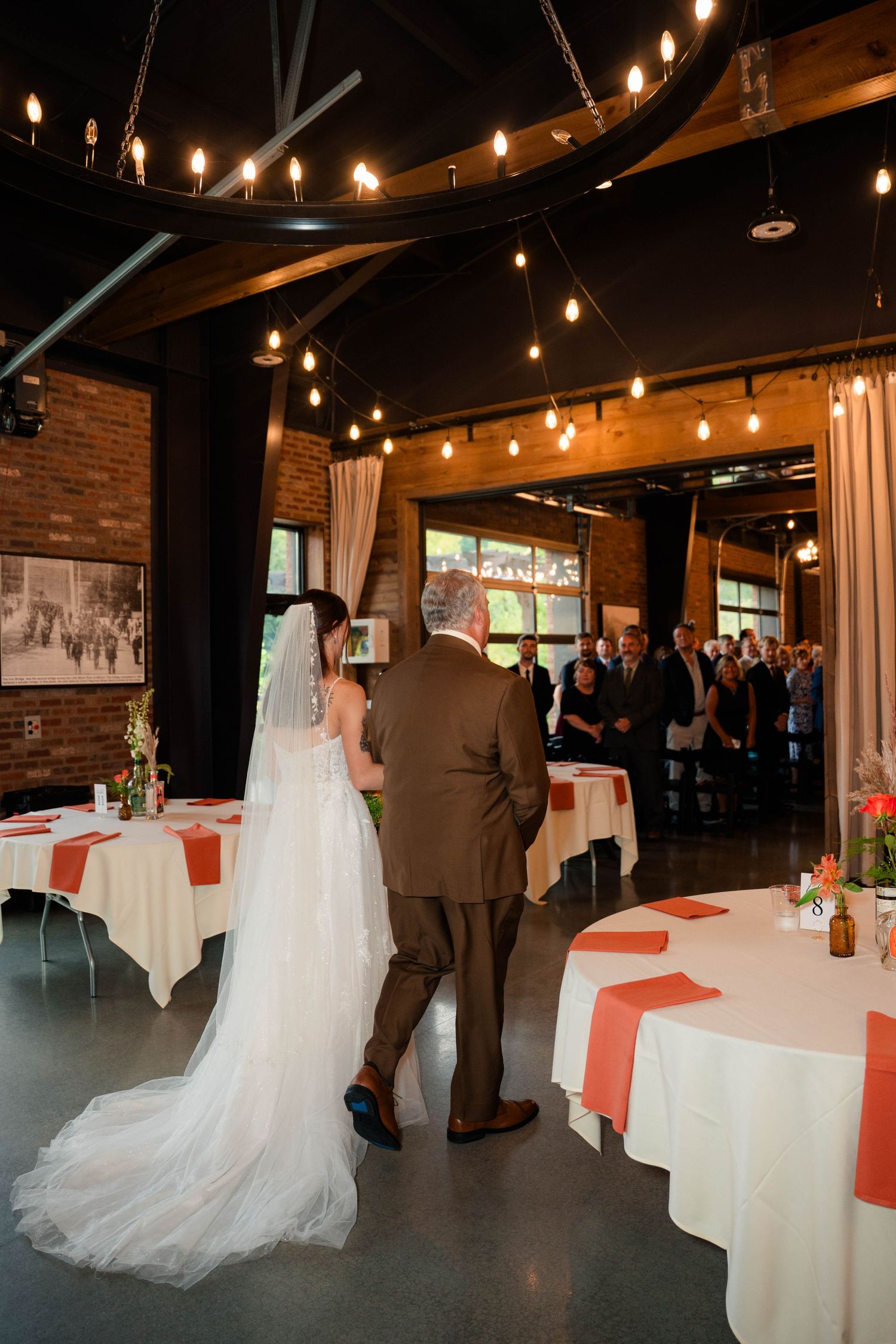 Bride walks down the aisle with escort through elegantly lit industrial wedding venue with string lights.