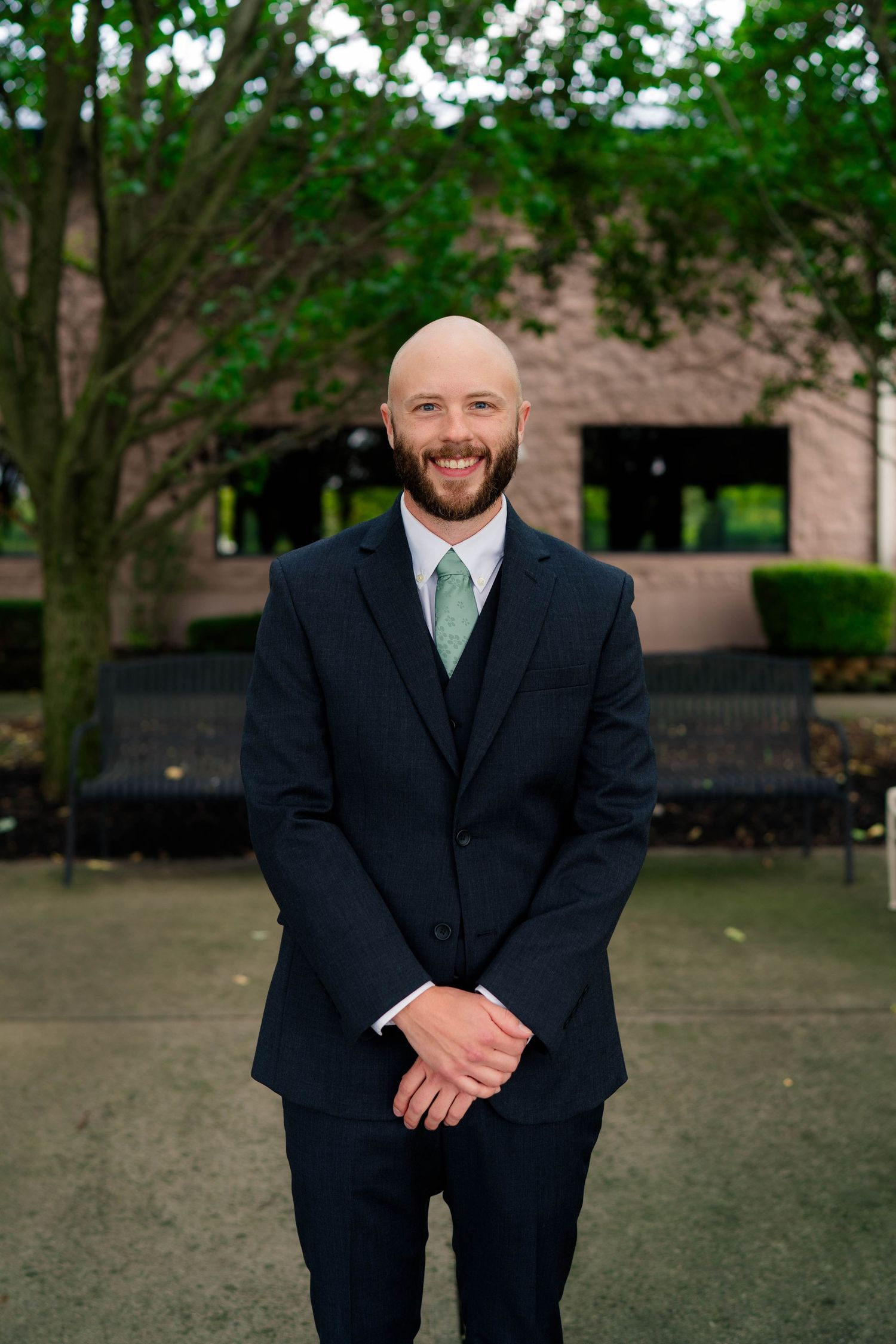 Smiling bald groom with beard in navy three-piece suit and mint green tie standing outdoors before wedding ceremony.