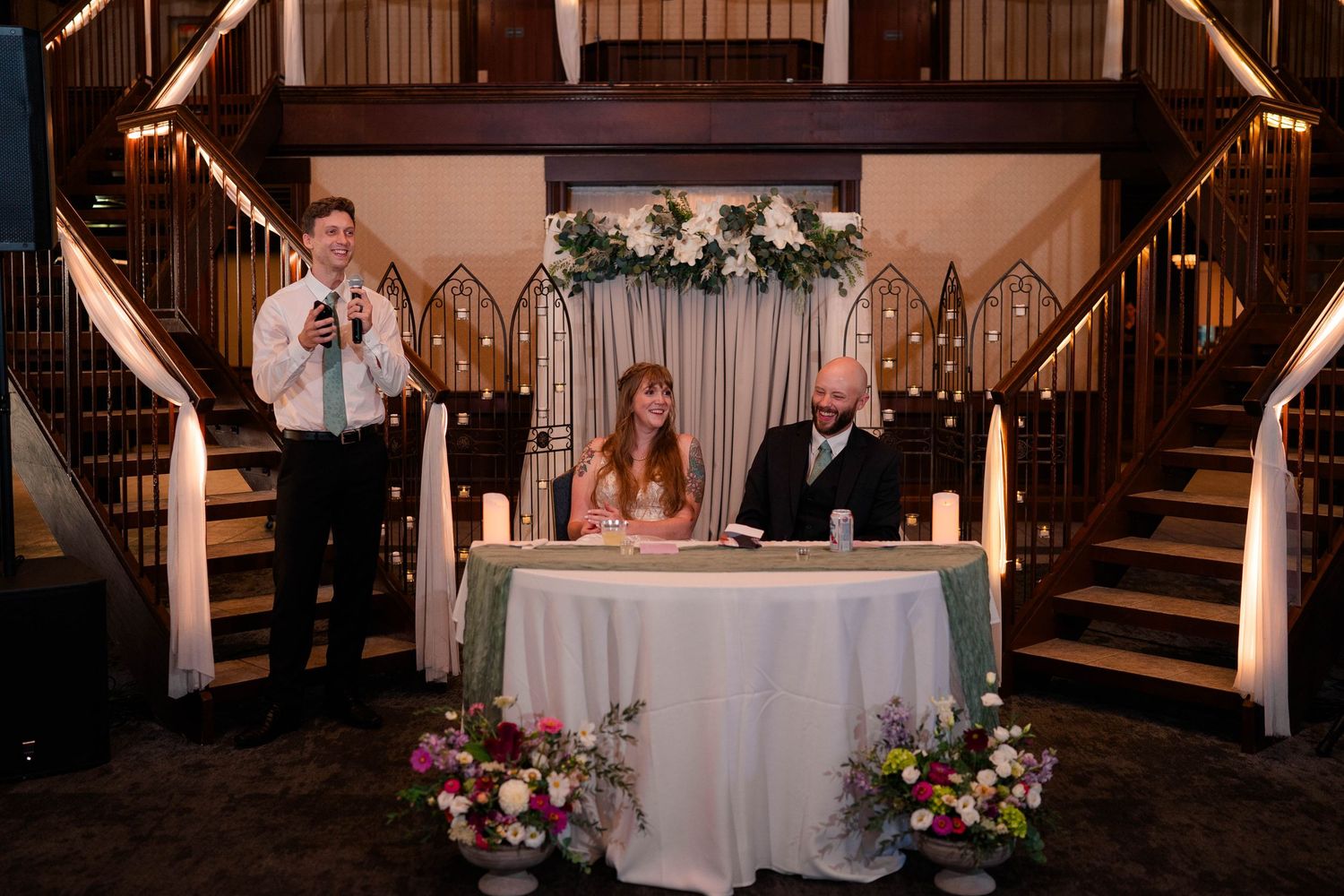Bride and groom sit at sweetheart table with candles during indoor wedding reception ceremony signing.