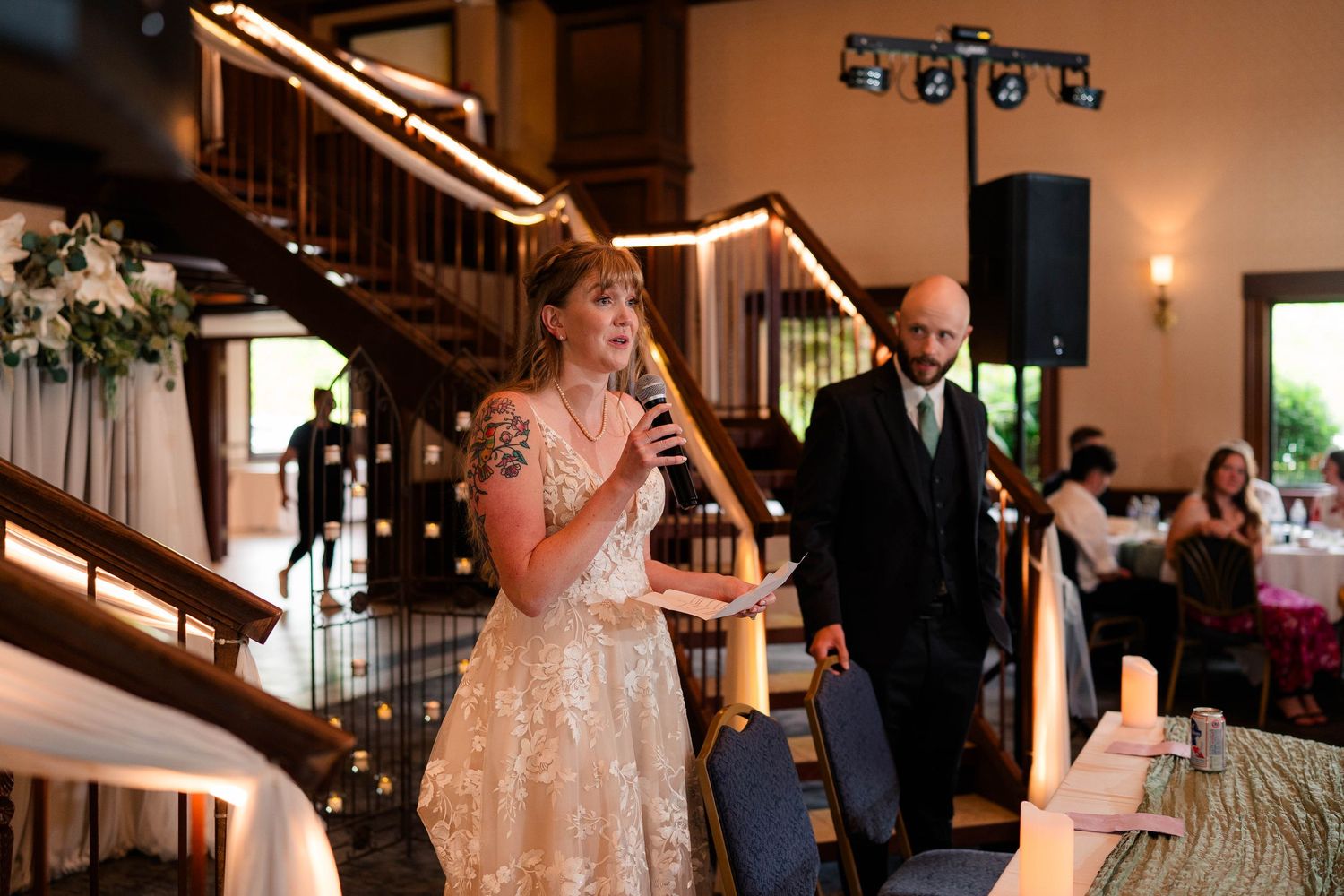Bride in a lace gown speaks into a microphone at the wedding reception while groom stands proudly behind her.