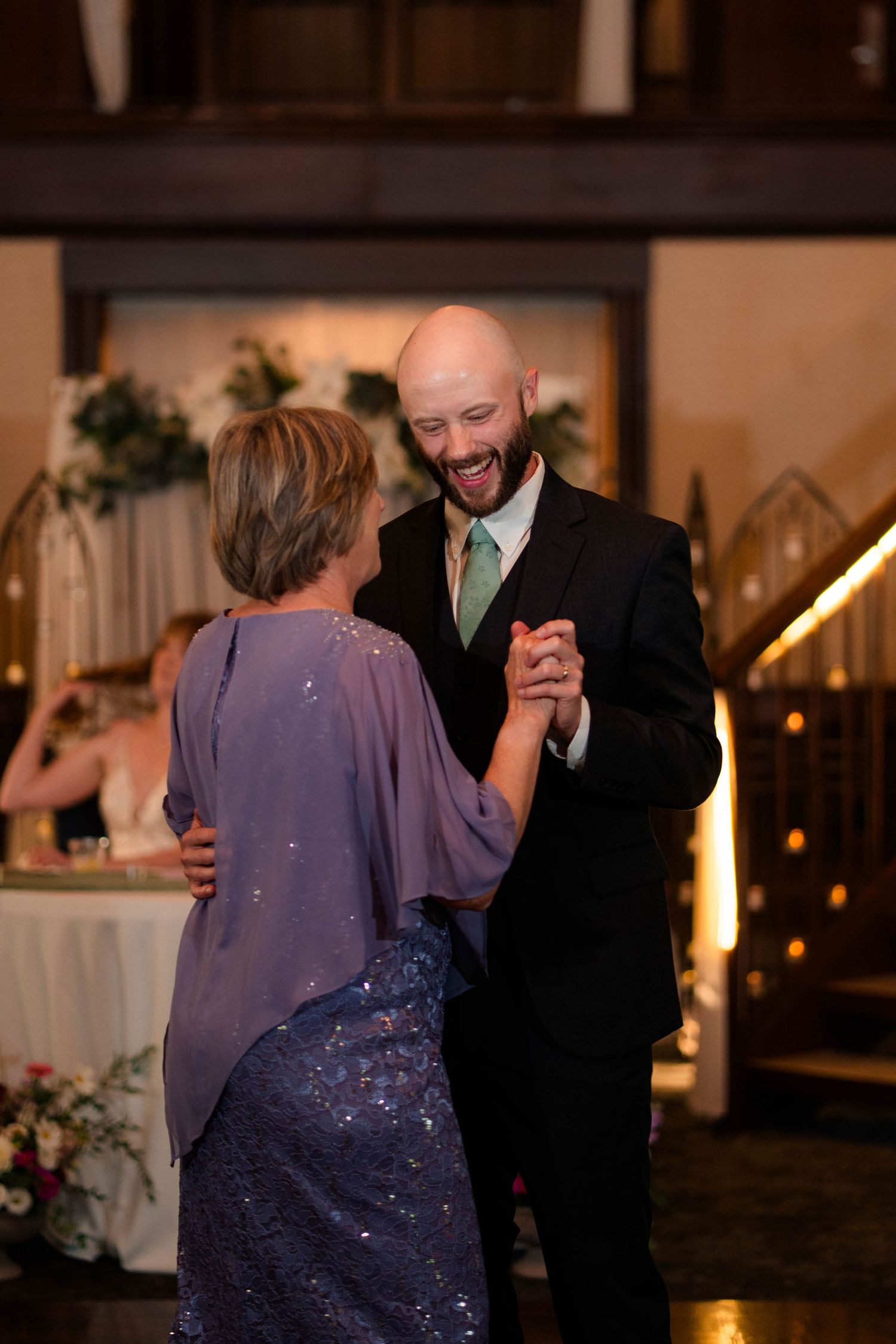 Smiling bald groom dances with mother in purple sequin dress at wedding reception.