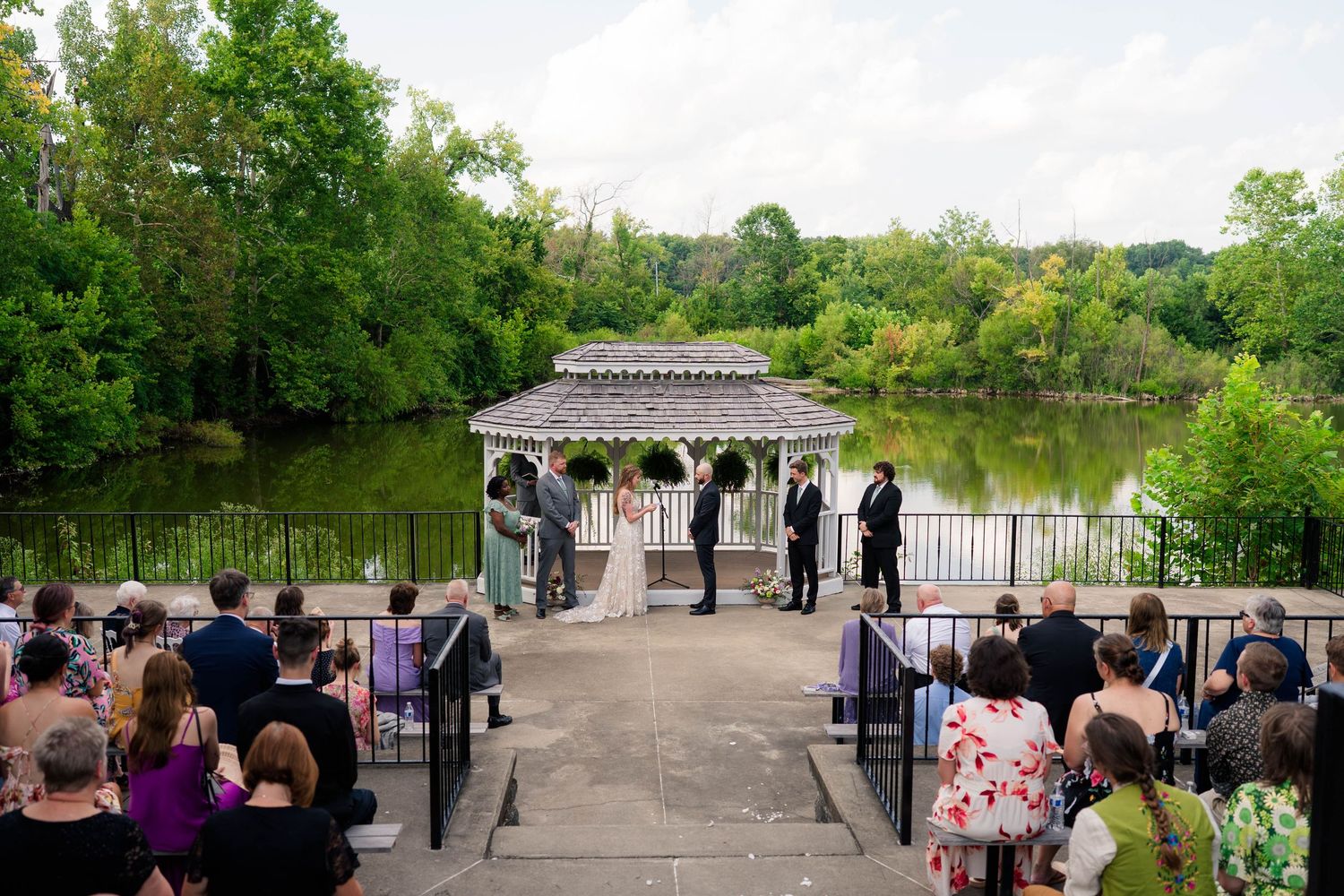 Outdoor wedding ceremony at a gazebo overlooking a serene lake surrounded by lush green trees.