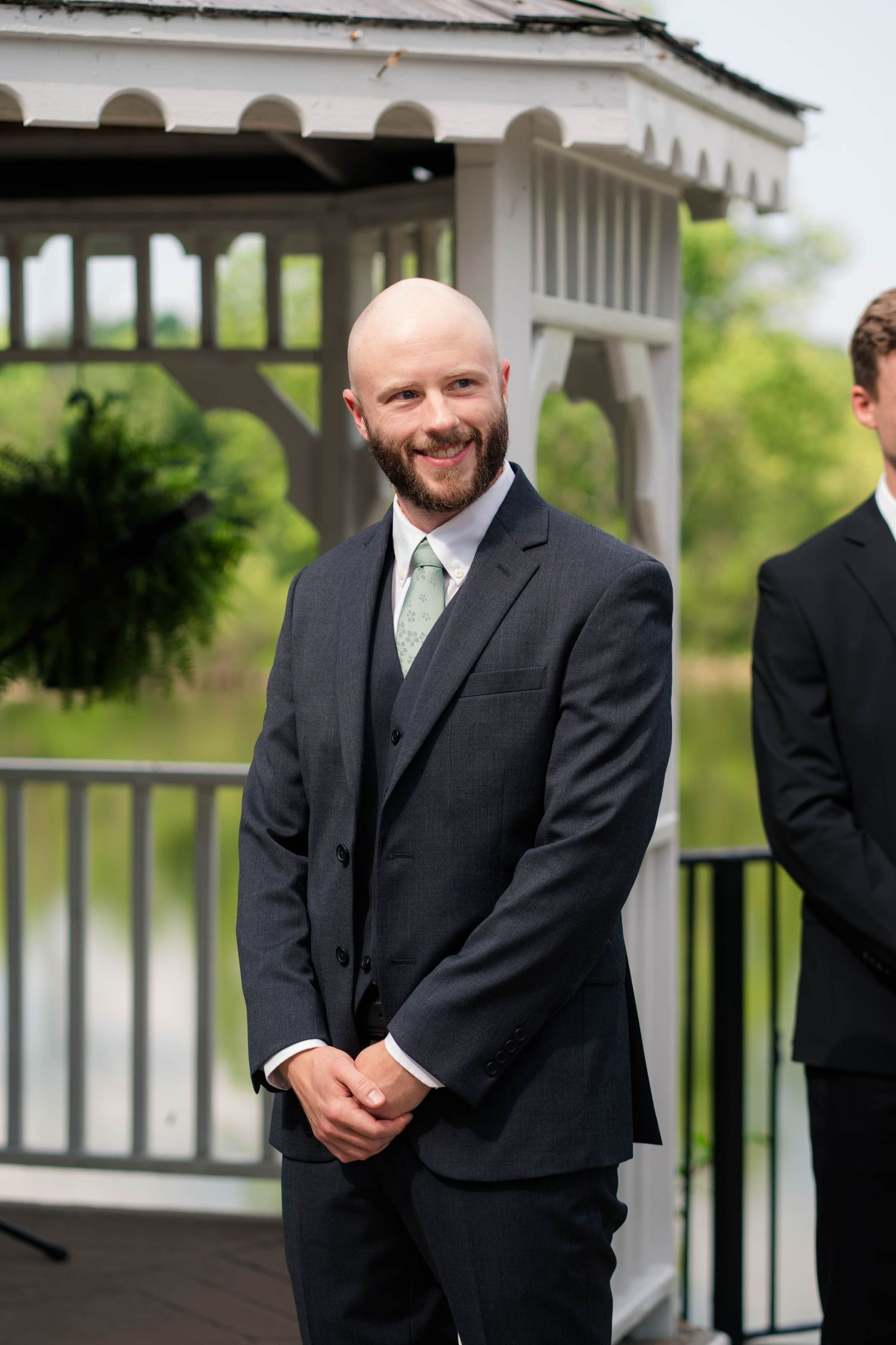 Smiling bald groom in dark suit with green tie stands at outdoor wedding ceremony near gazebo.