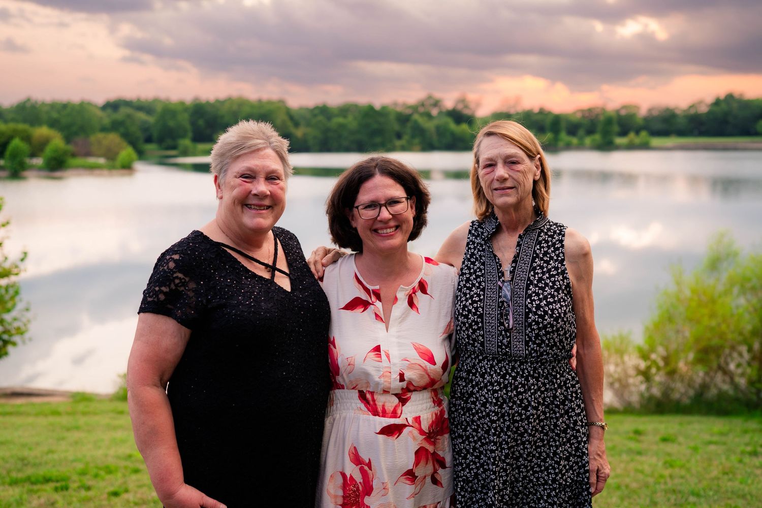 Three women smile together near a scenic lake at sunset during outdoor wedding celebration.