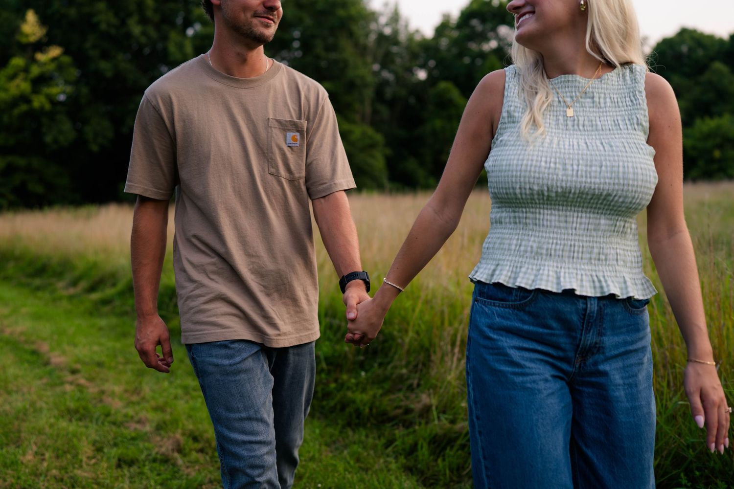 A couple walks hand in hand through a green meadow, woman smiling in a light blue smocked top and jeans.