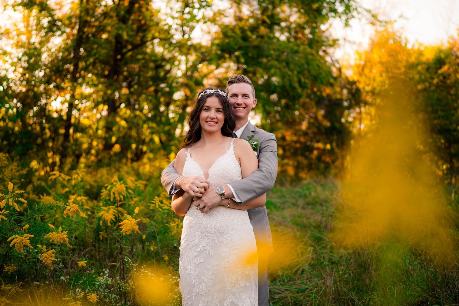 Happy bride and groom embrace surrounded by golden wildflowers in warm sunset light.
