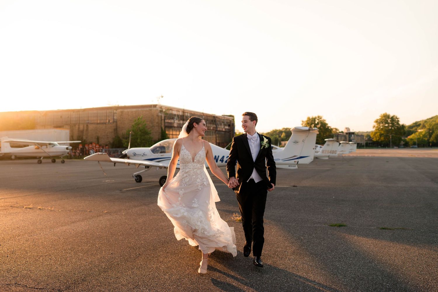 Bride and groom walking hand in hand on airport tarmac at golden sunset with planes behind them.