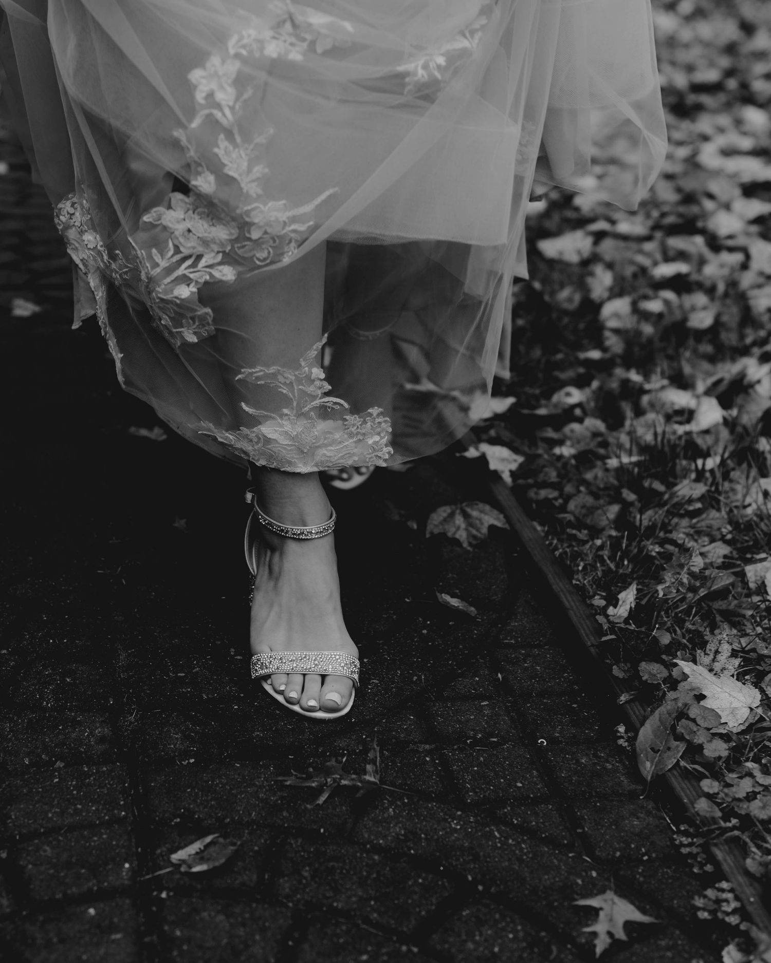 Black and white close-up of bride's strappy heeled sandals beneath a floral lace wedding gown.